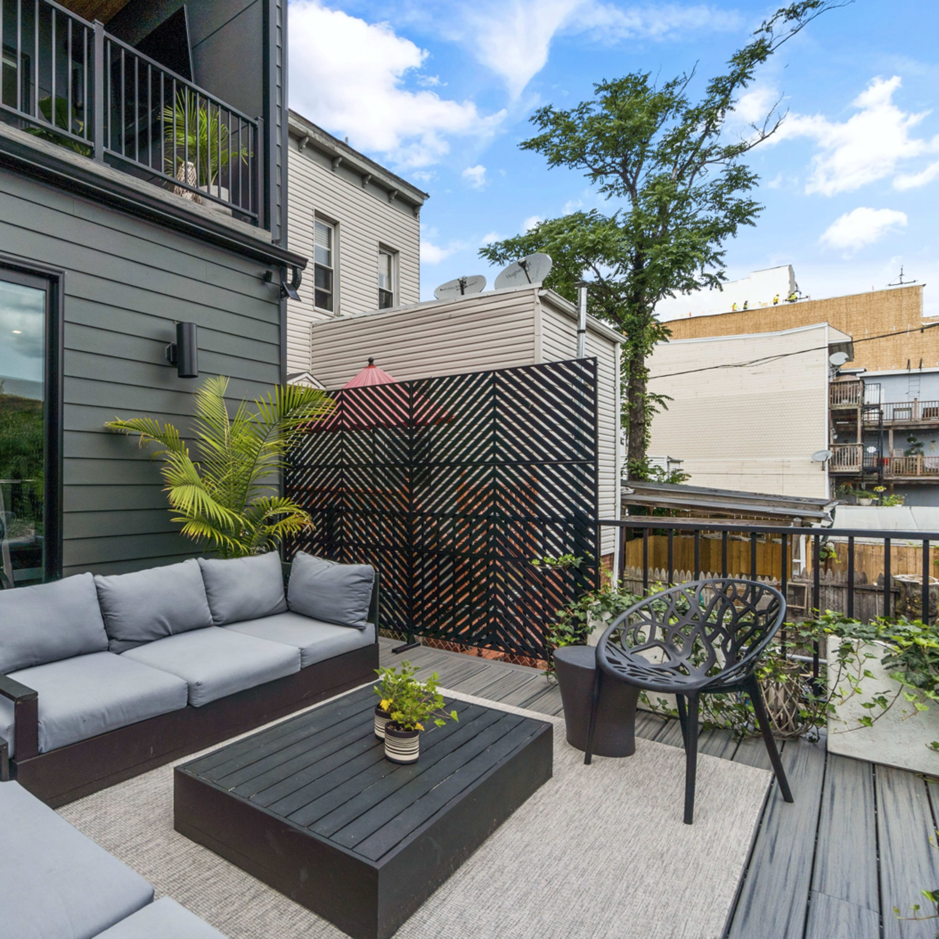 A modern outdoor patio features gray seating, a black table, and a decorative black screen, surrounded by greenery and urban buildings.