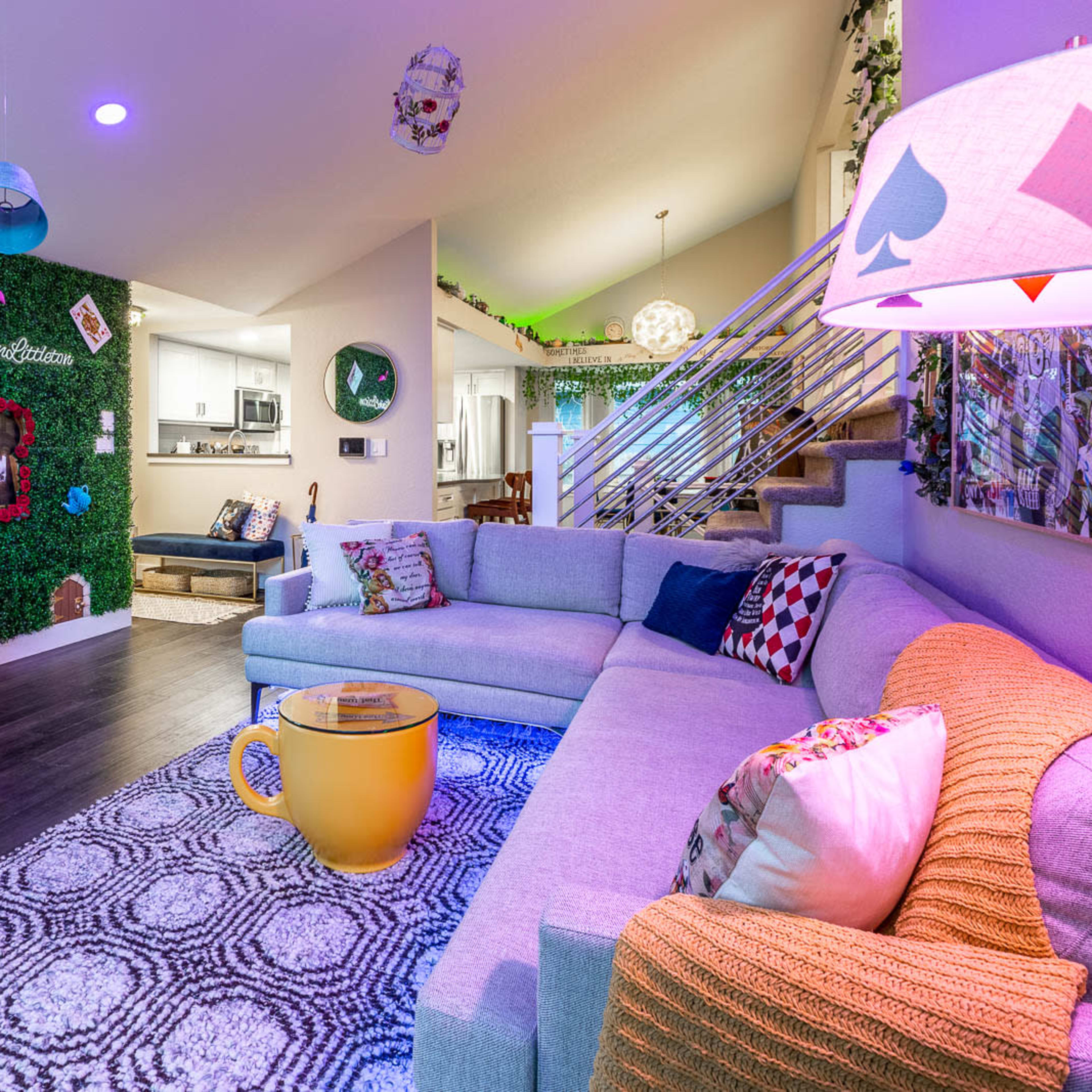 A modern living room features a gray sectional sofa, a brown leather chair, and decorative elements like potted plants and a large yellow cup-shaped table.