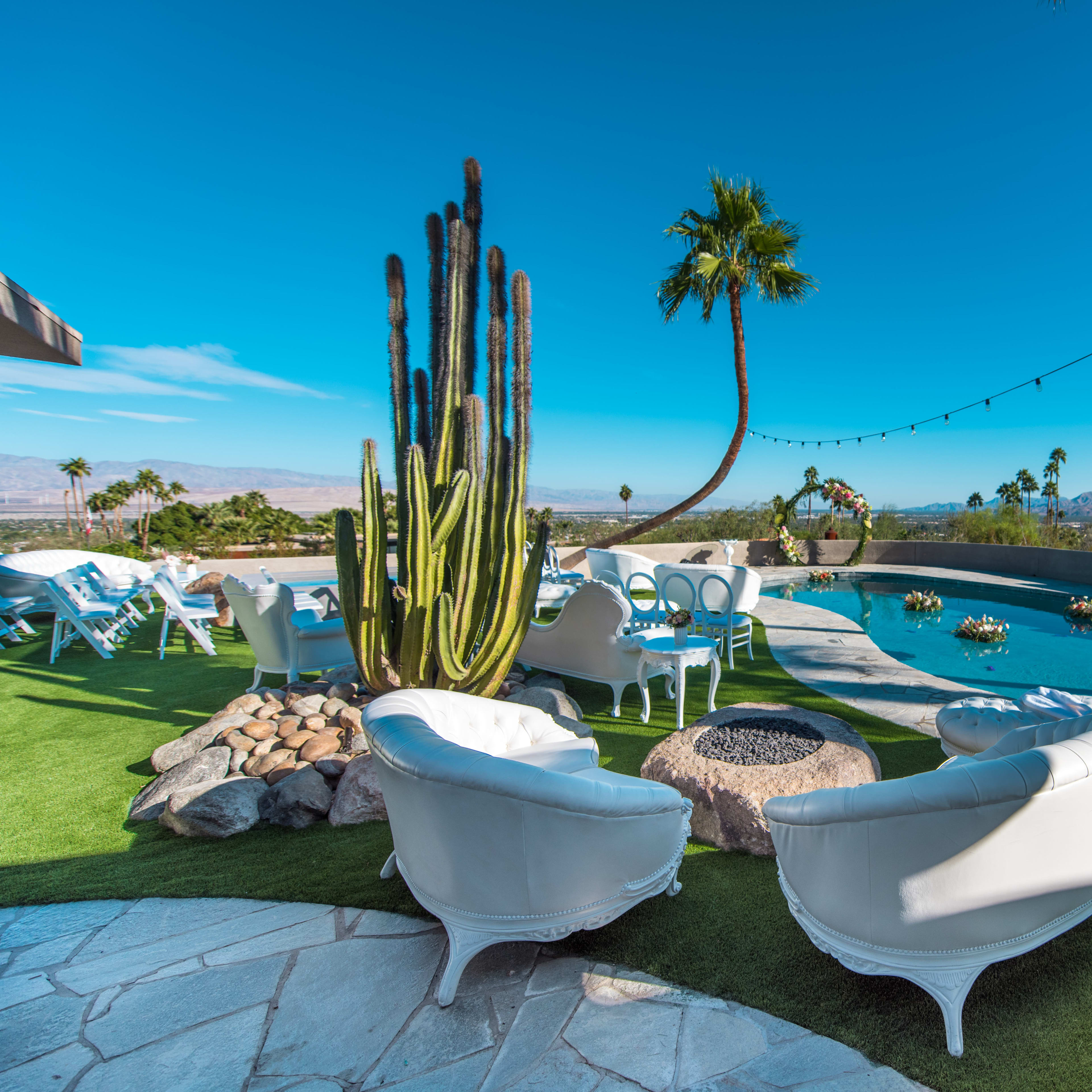 A patio area with white outdoor furniture, a swimming pool, and a large cactus, surrounded by palm trees and mountains in the background.