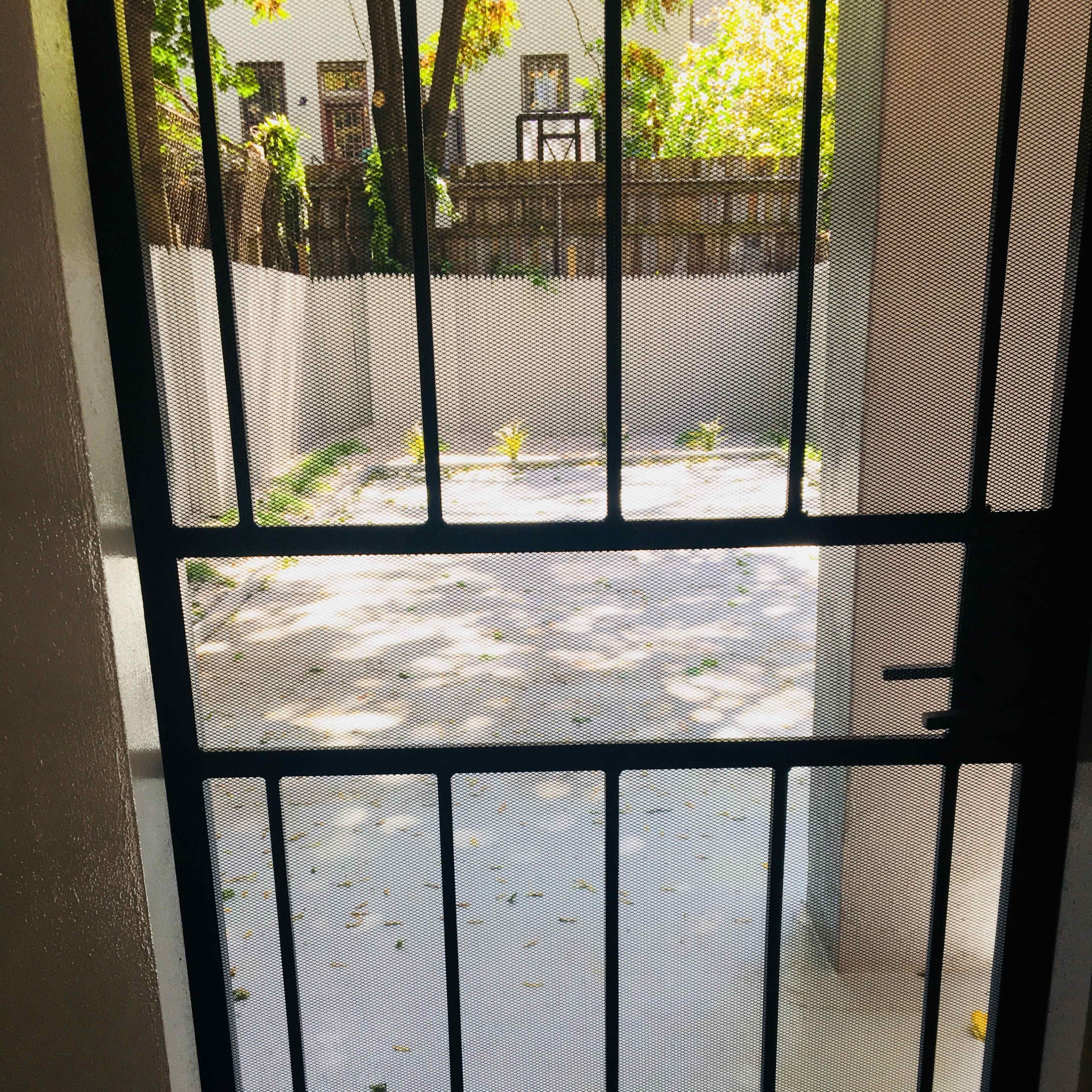 A black metal gate opens to a gravel courtyard surrounded by concrete walls and shaded by trees.
