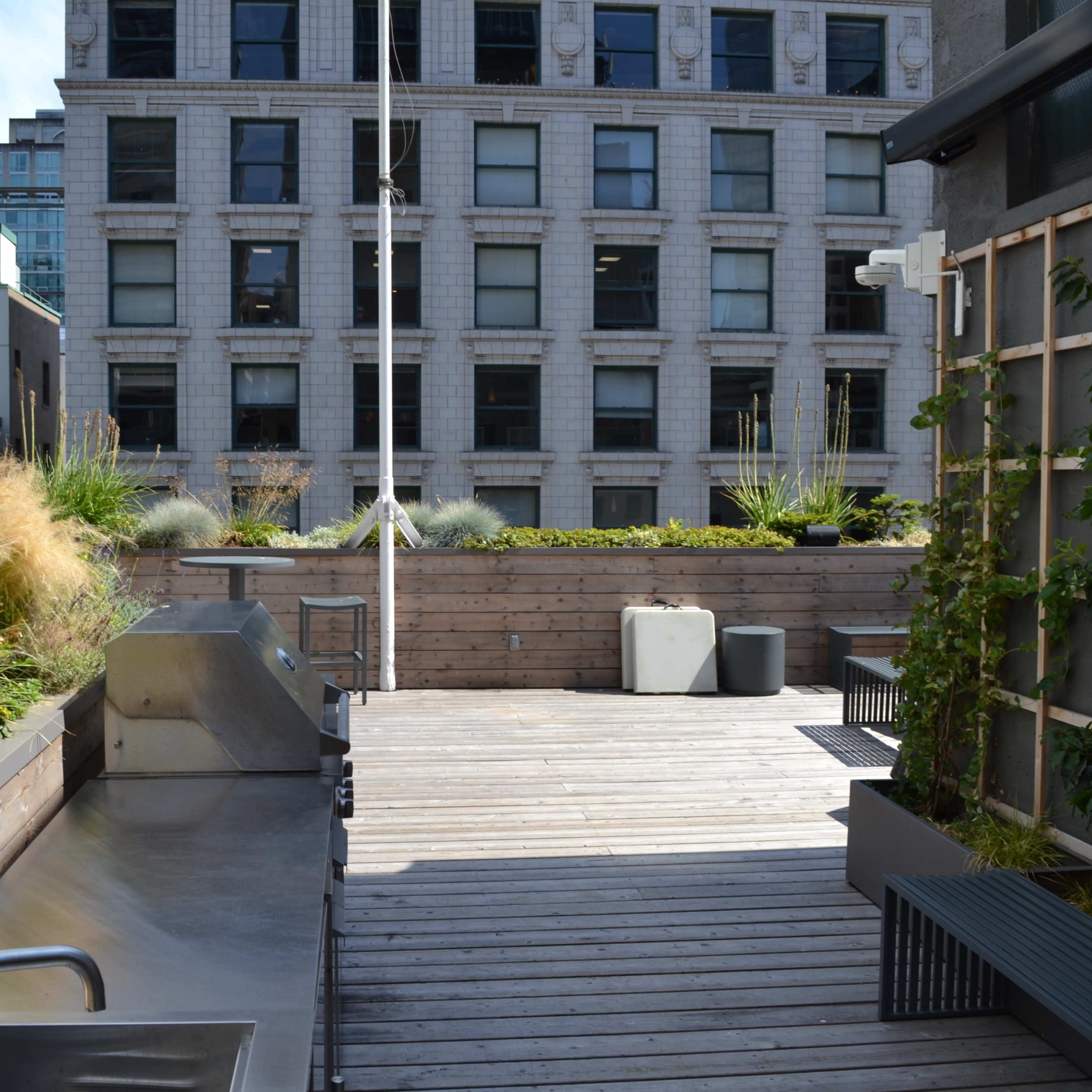A rooftop terrace with wooden flooring, a stainless steel grill, and planters filled with greenery, overlooking a neighboring building.