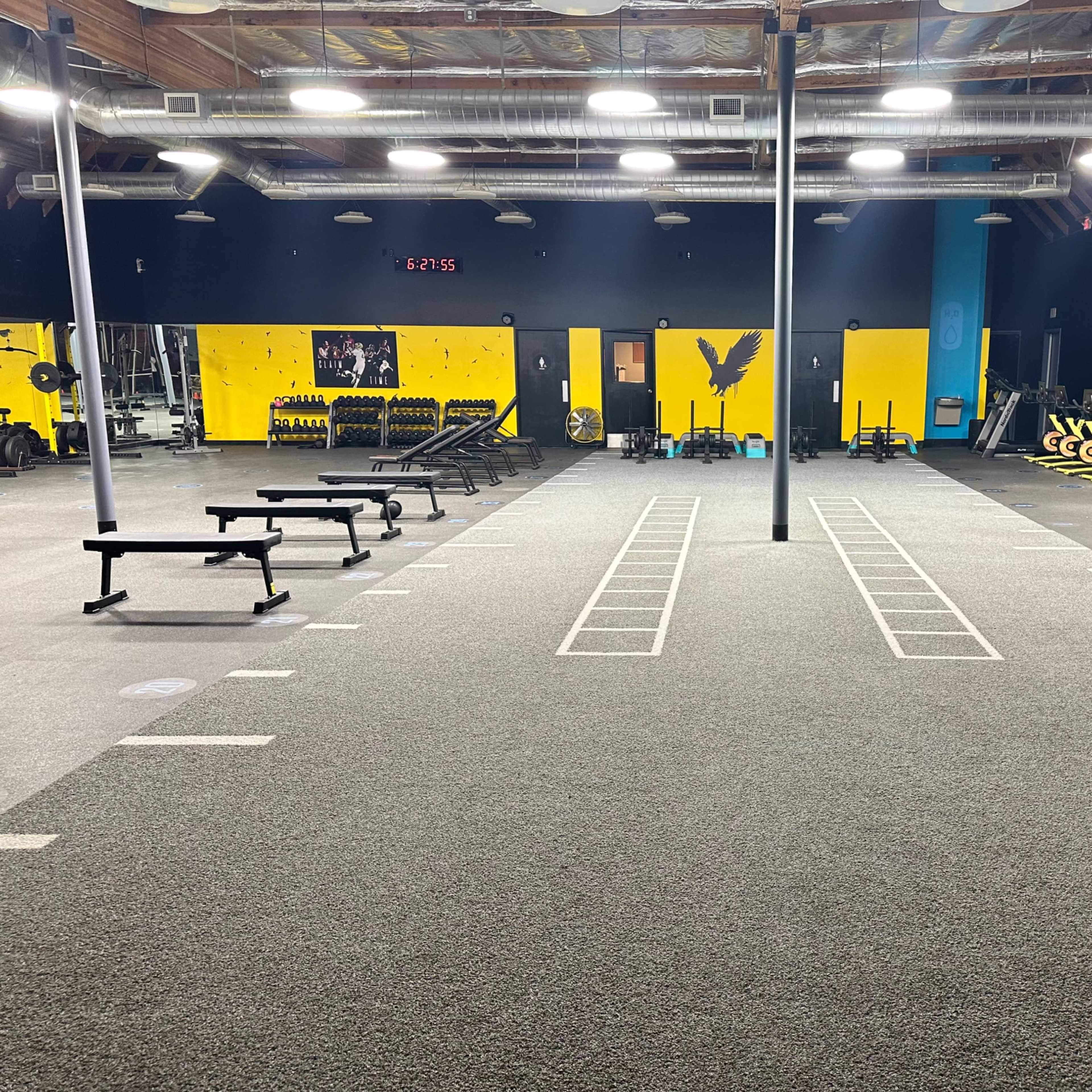 A spacious gym with workout equipment, including benches, weights, and exercise bikes, arranged in a well-lit area with a dark wall and yellow accents.