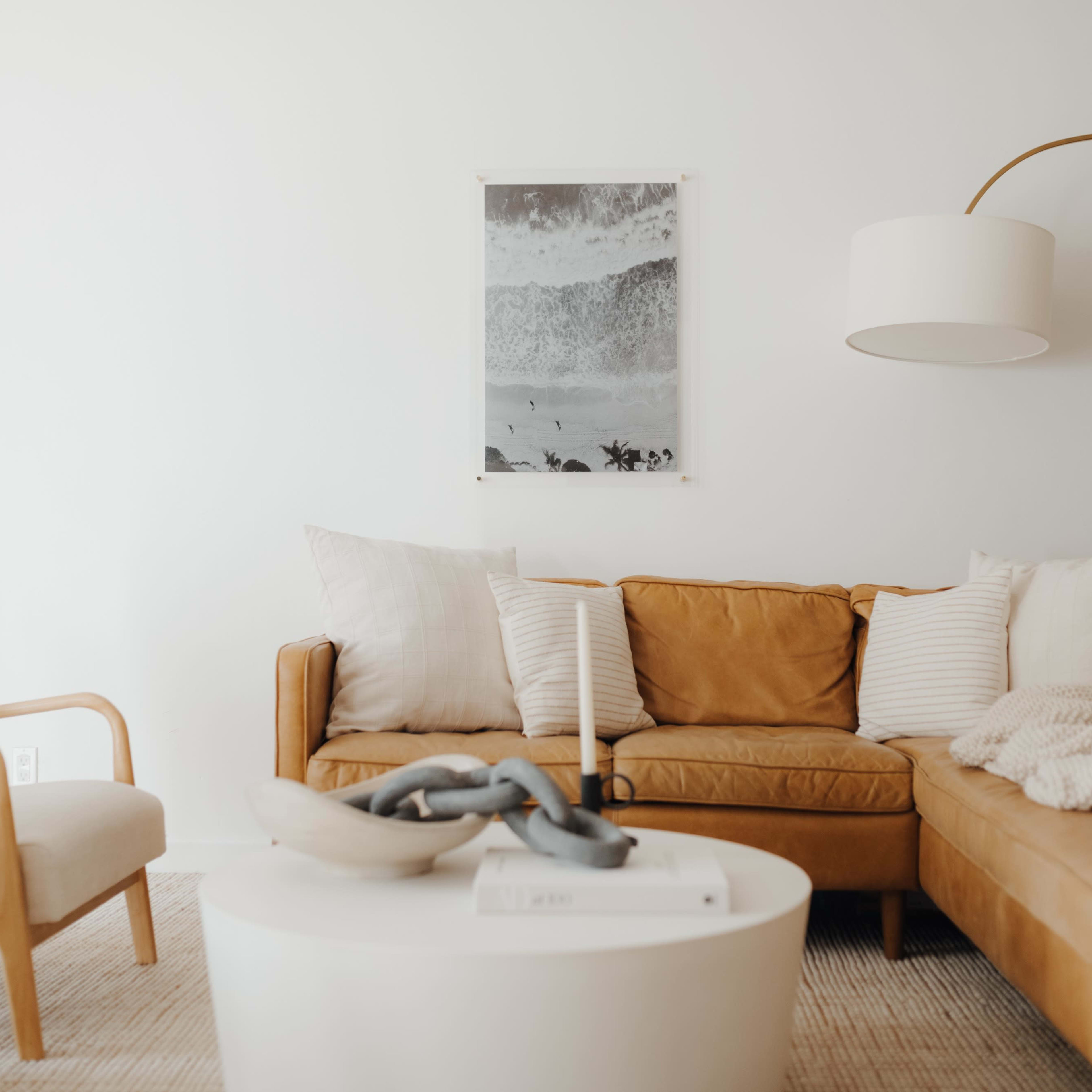 A living room features a brown leather sofa with decorative pillows, a round white coffee table, and a floor lamp, all set against a white wall adorned with a framed artwork.