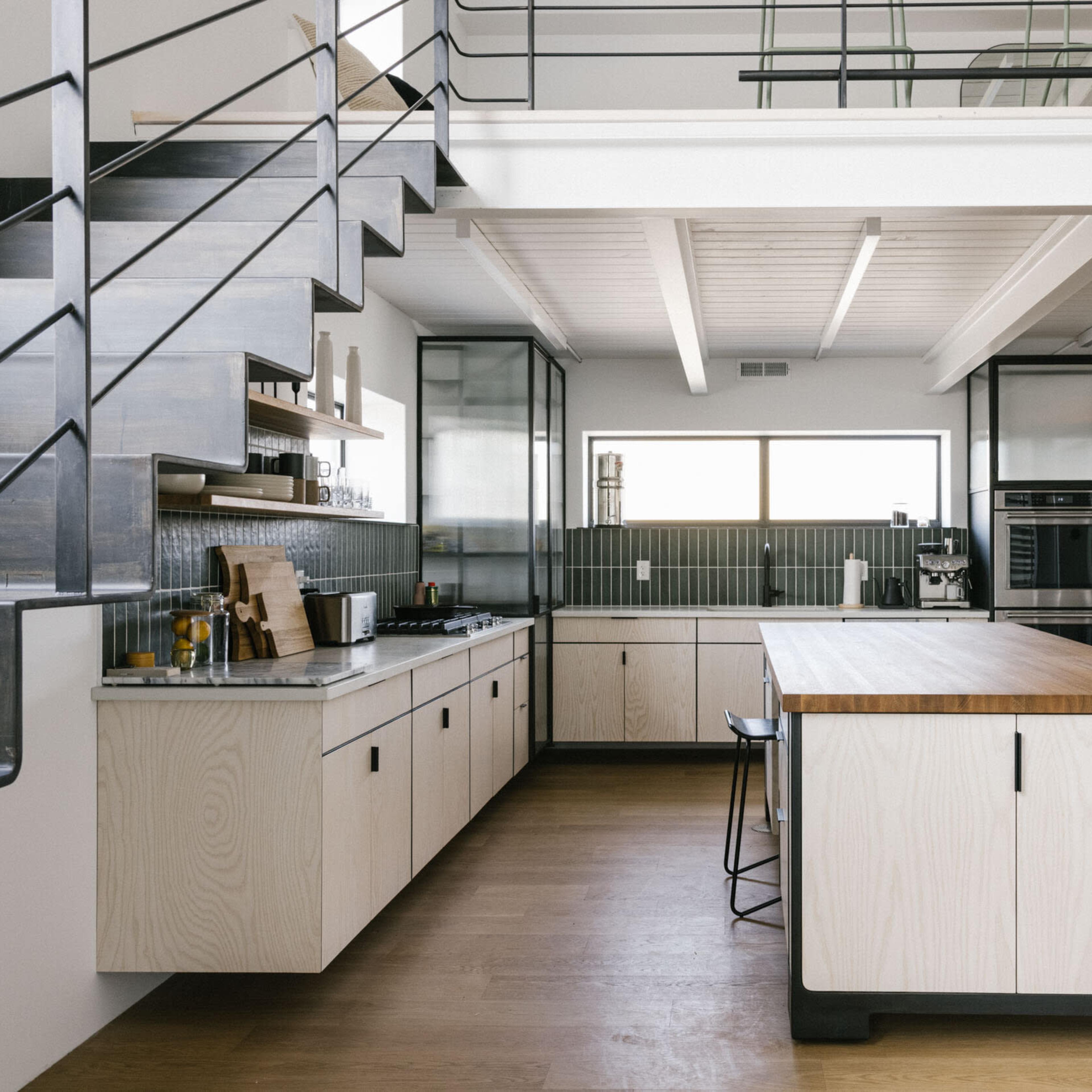 A modern kitchen features a central island with a wooden countertop, sleek cabinetry, stainless steel appliances, and a staircase leading to an upper level.