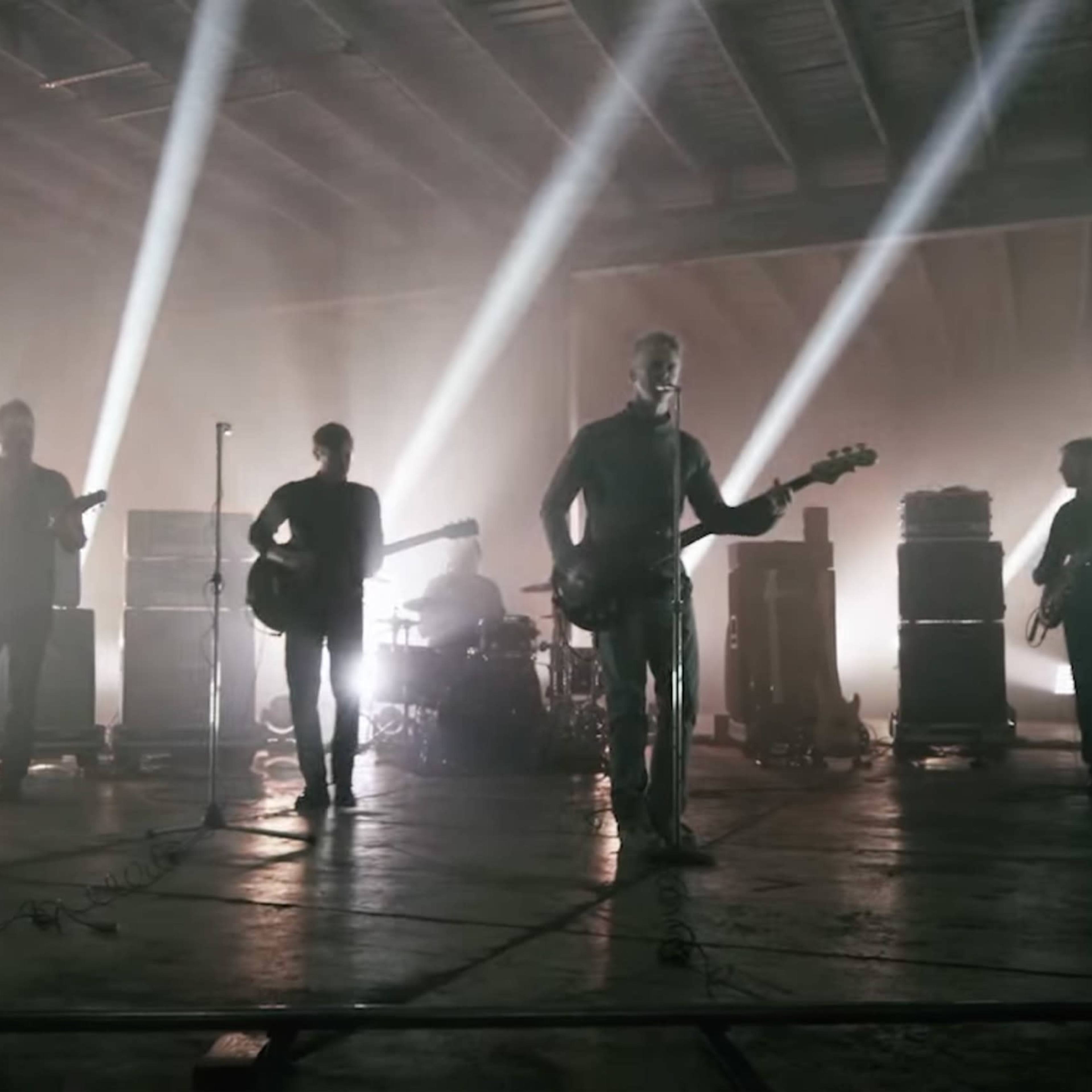 A band performs onstage in a dimly lit warehouse with bright beams of light illuminating the musicians from behind.