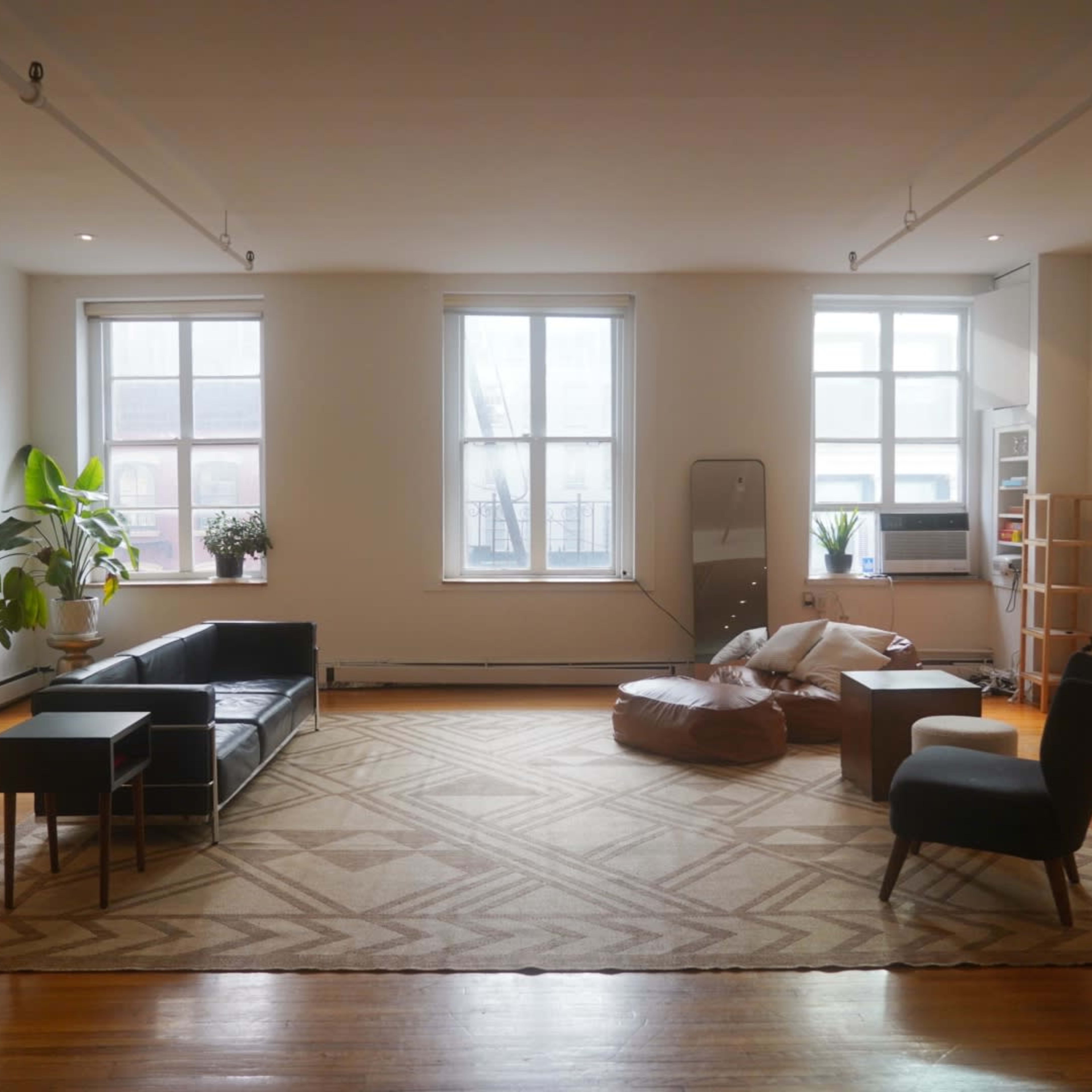 A spacious living room with a dark couch, a coffee table, a beanbag chair, and large windows letting in natural light.
