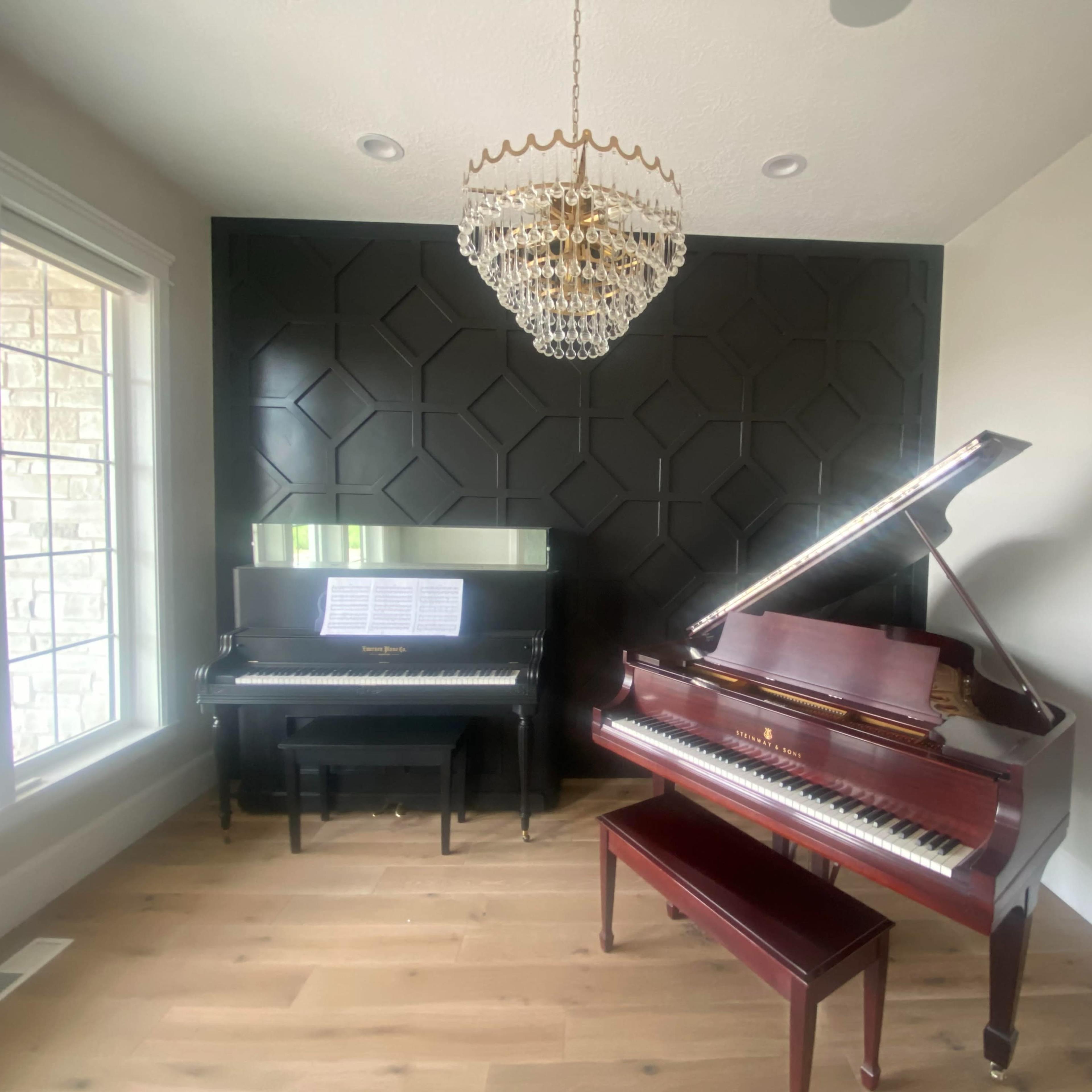 A room with two pianos, a grand piano on the right and an upright piano on the left, accompanied by a chandelier overhead.