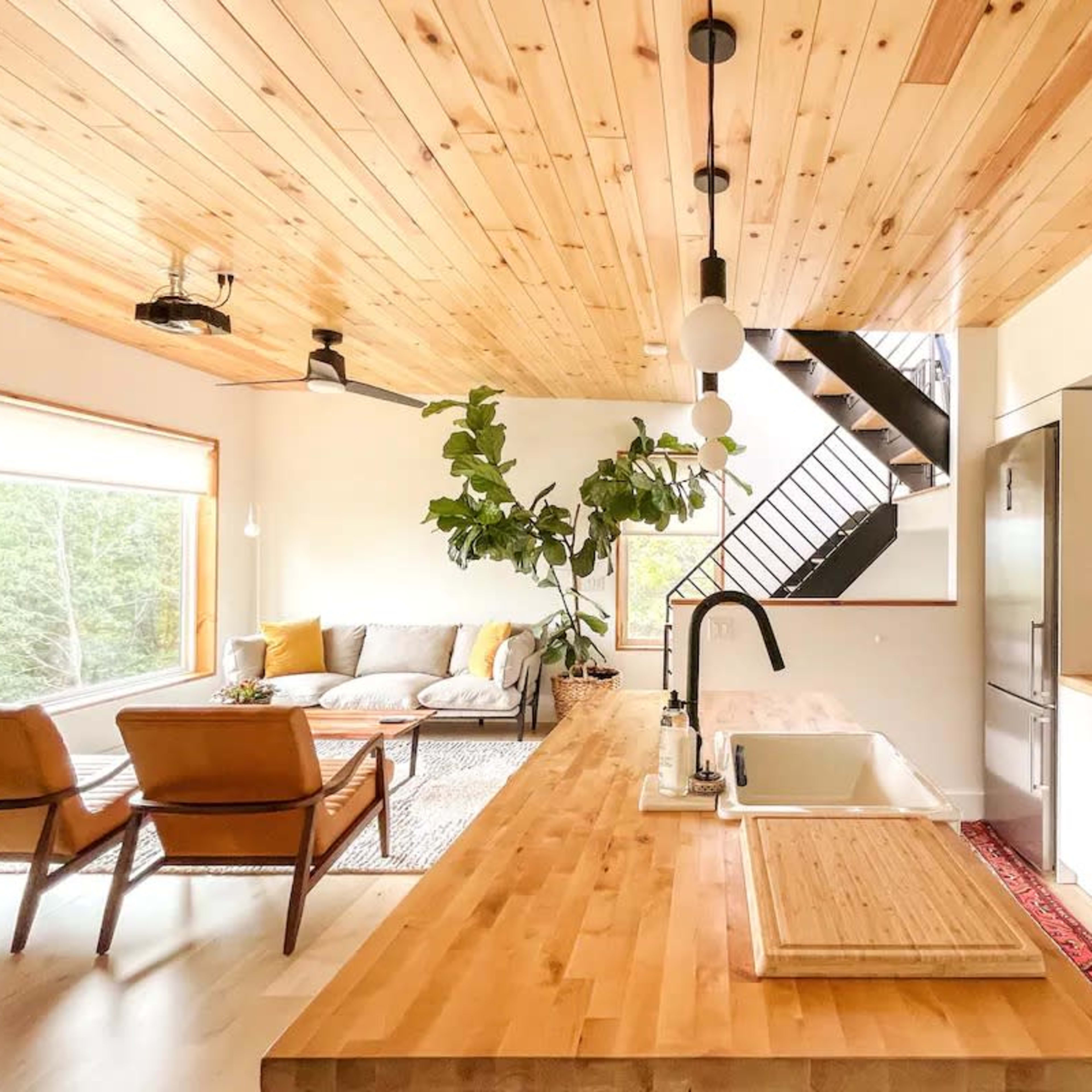 A modern kitchen with wooden ceilings, a large island with a sink, and an open living area featuring a sofa and large windows.