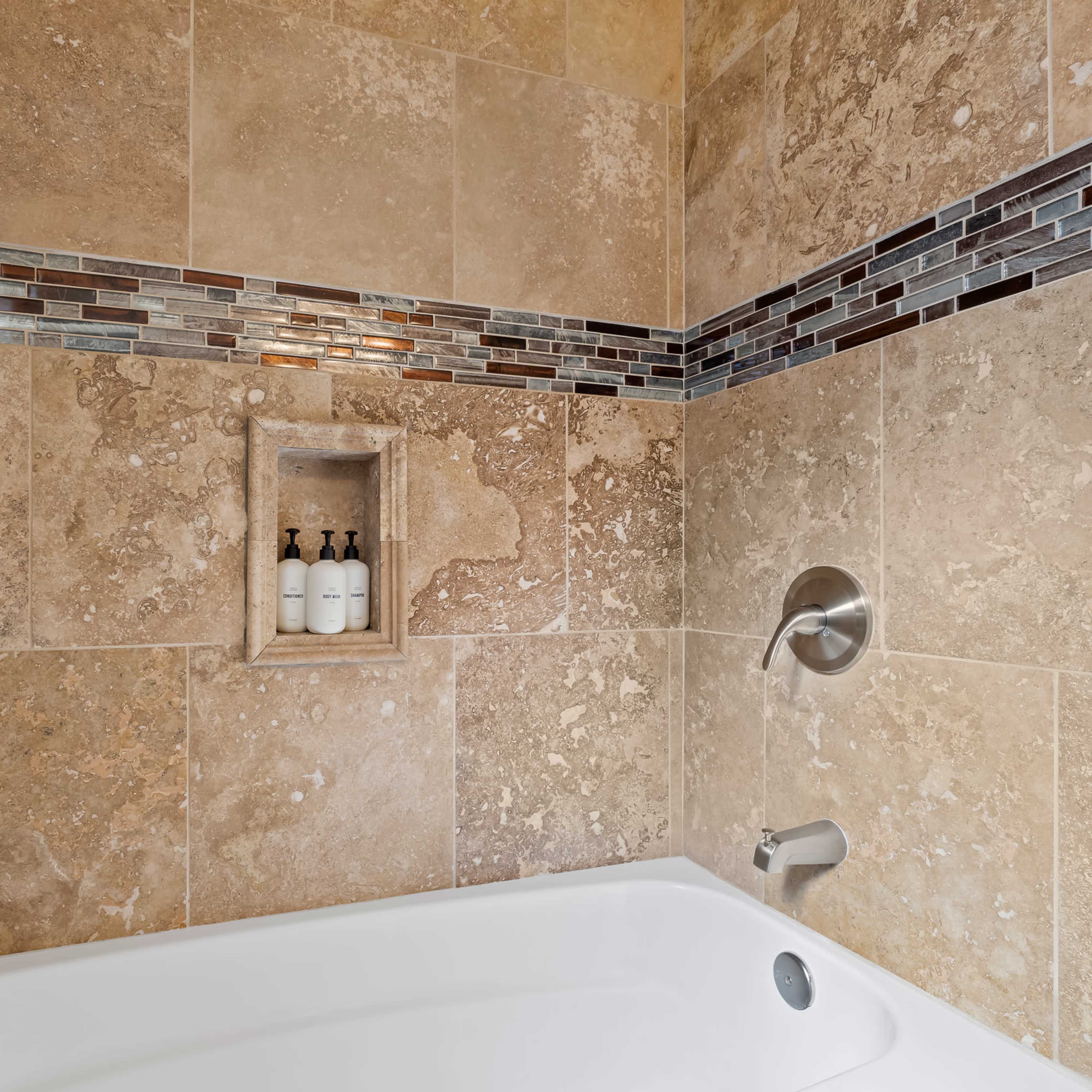 A tiled shower with a built-in shelf holding three bottles, featuring a tub at the base.