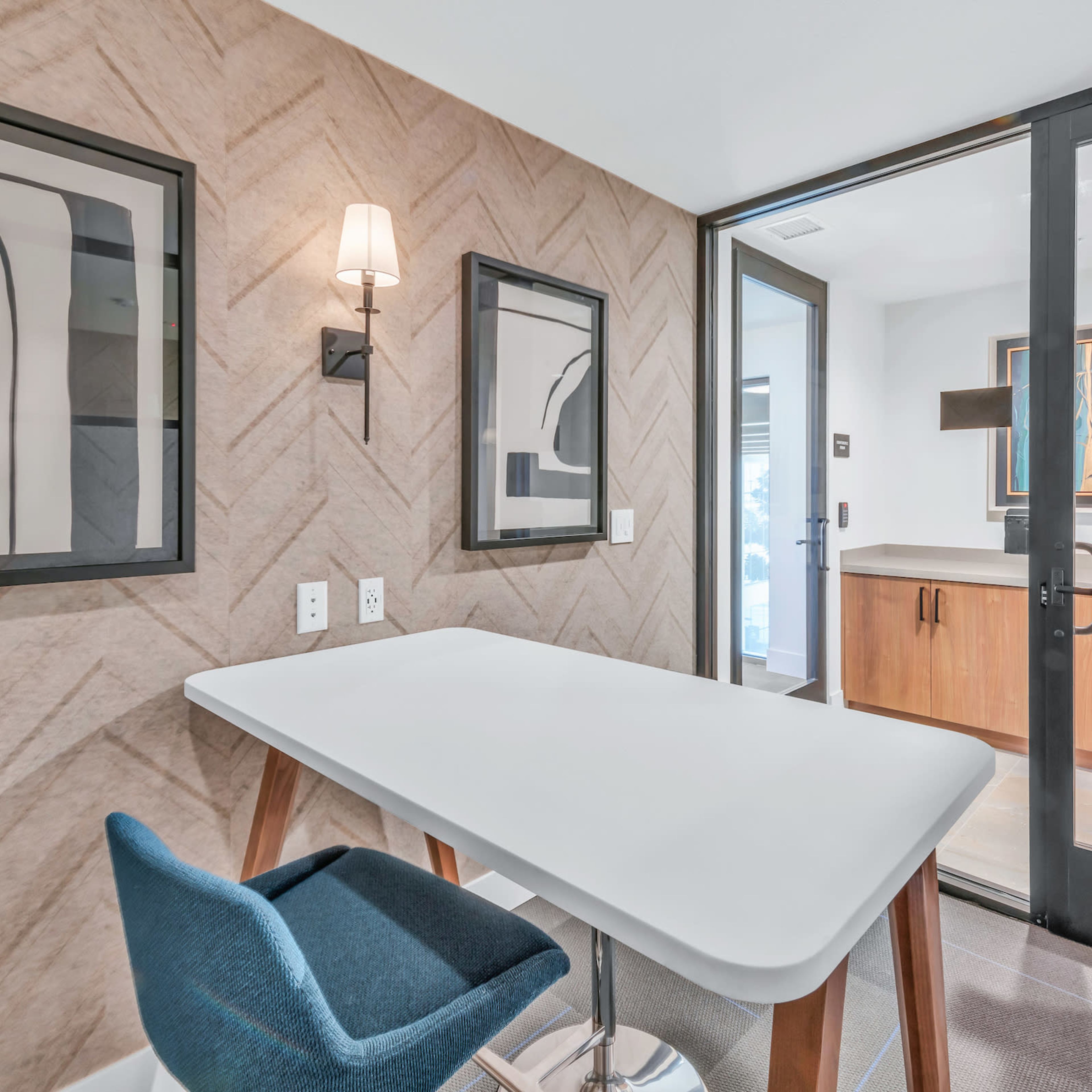 A modern office space features a minimalist white table, a blue chair, framed abstract art on the walls, and a glass door leading to an adjacent area.