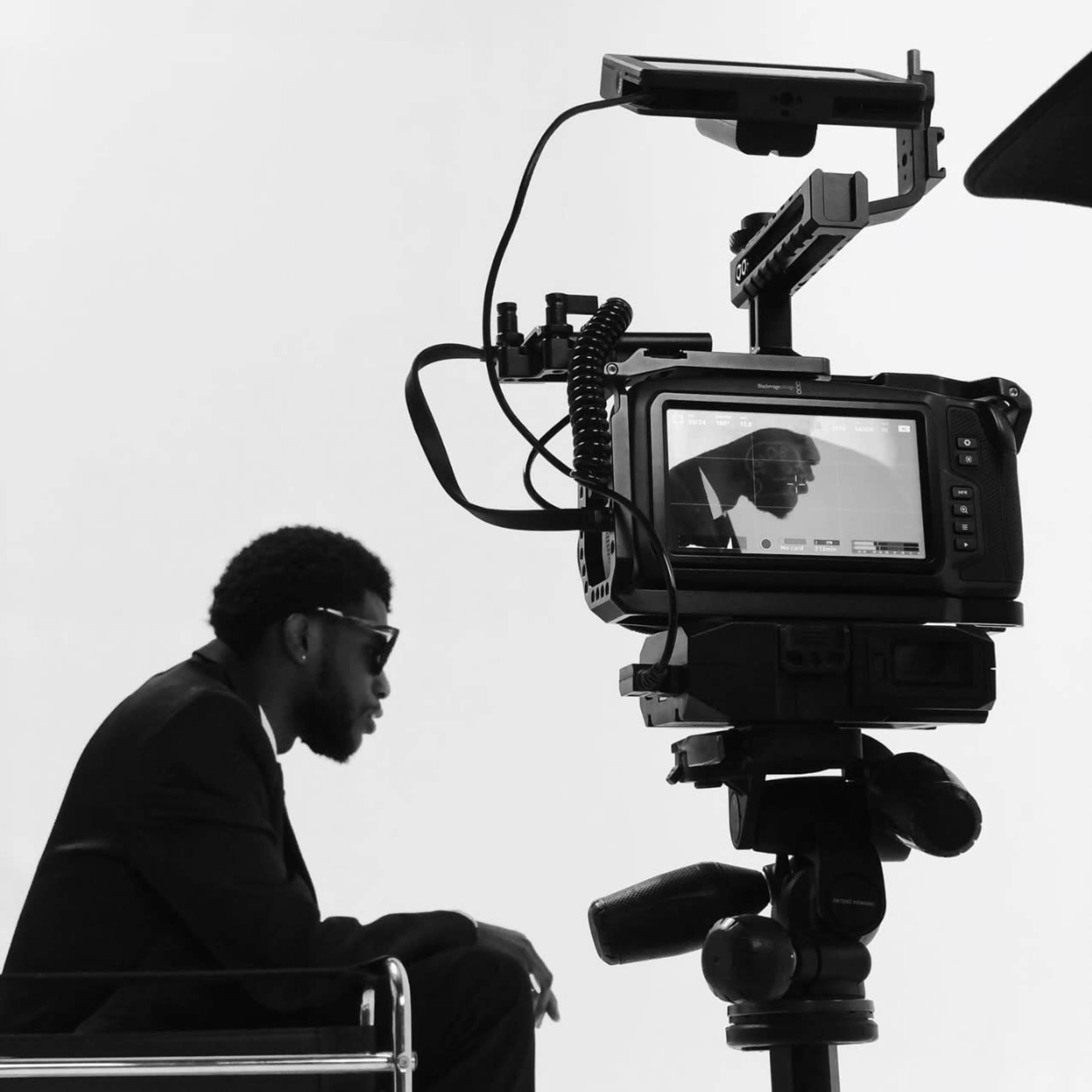 A man in a suit and sunglasses sits in profile, while a camera captures his reflection on its monitor.