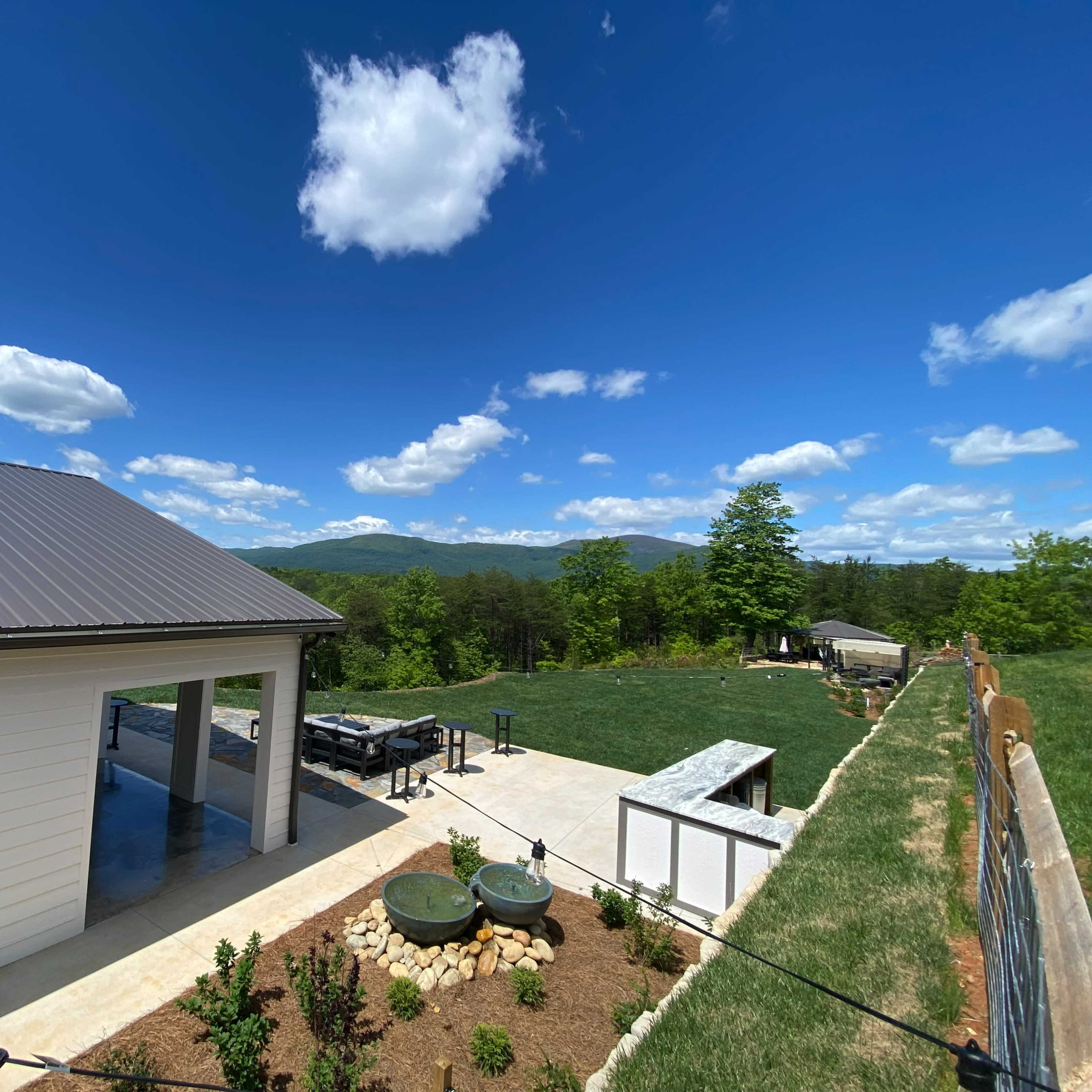 A spacious outdoor area features a modern building alongside a landscaped yard with mountains in the background under a blue sky with scattered clouds.
