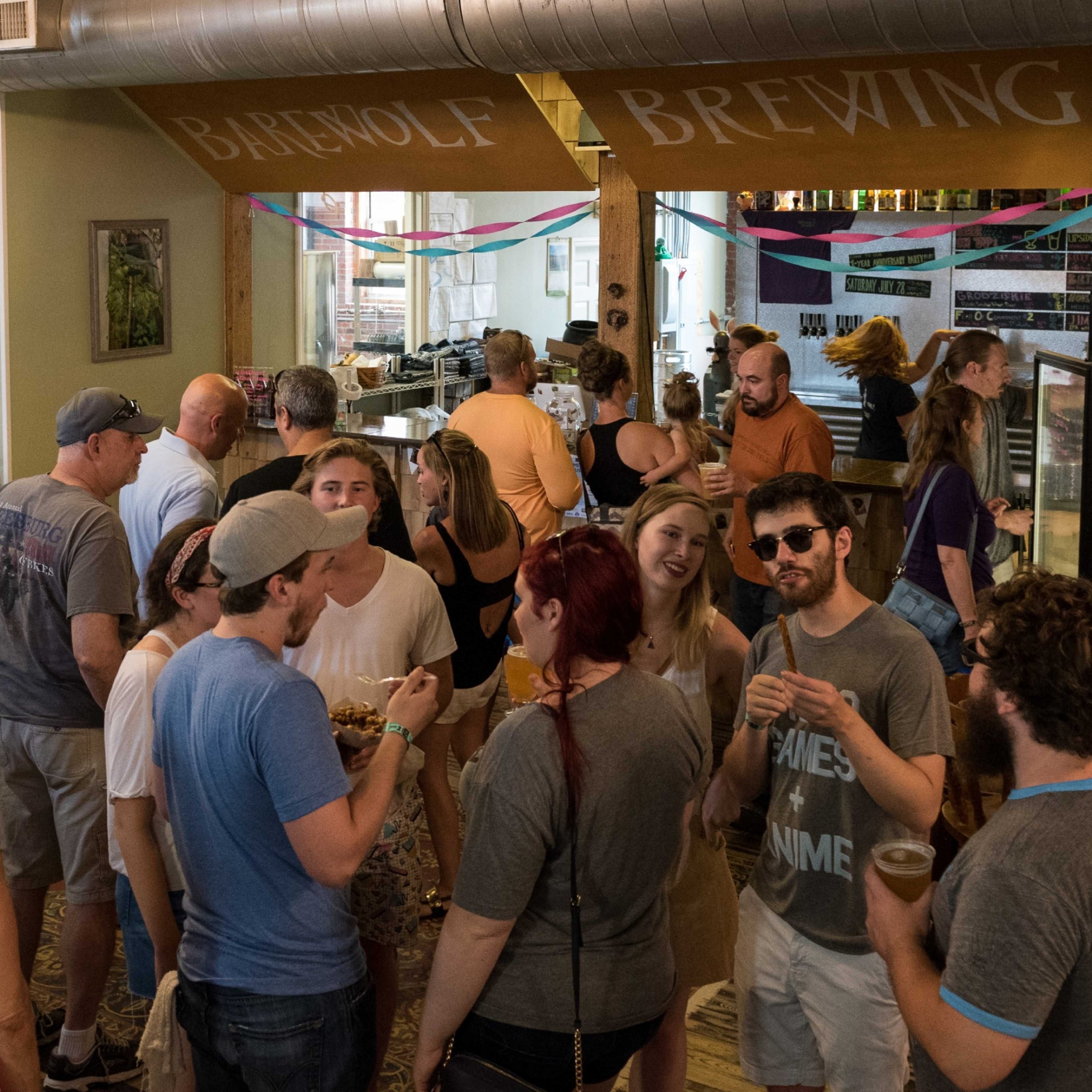 A crowd of people socializes inside a brewery, with some enjoying drinks and food at tables while others gather in clusters.