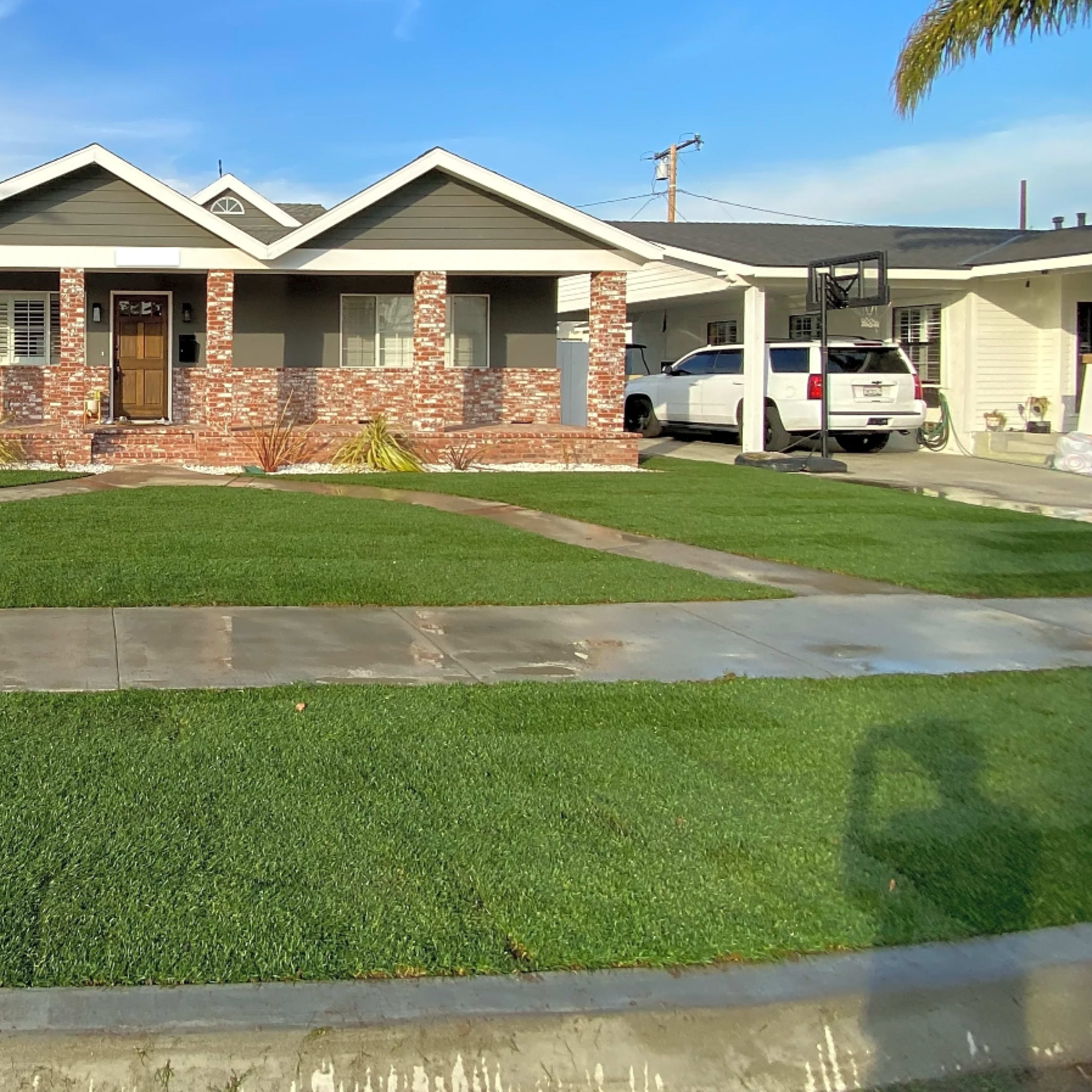 A single-story house with a brick facade, a landscaped front yard featuring green grass, and a driveway leading to a garage.