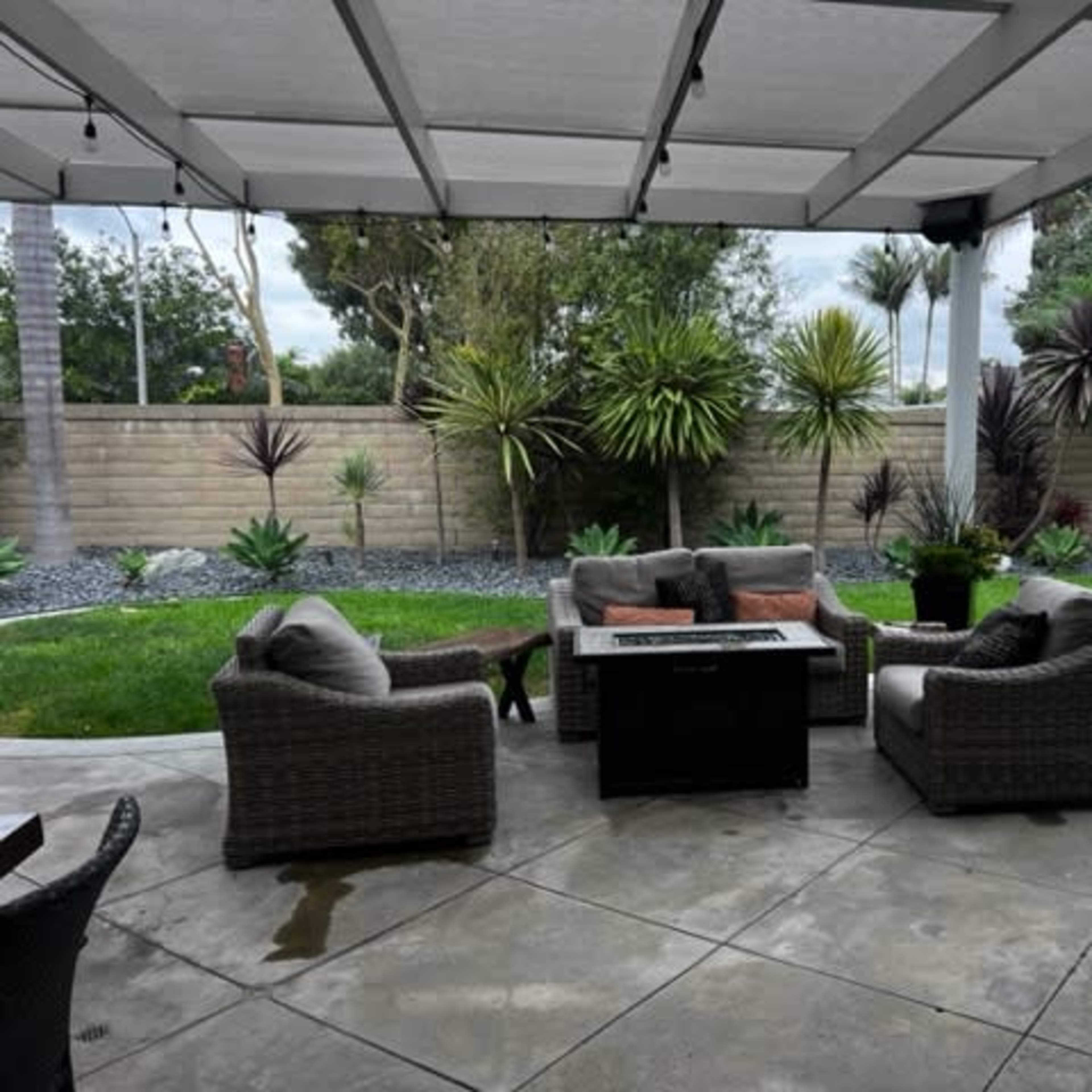 A covered patio features four wicker chairs arranged around a central table, with a landscaped garden of plants and grass in the background.