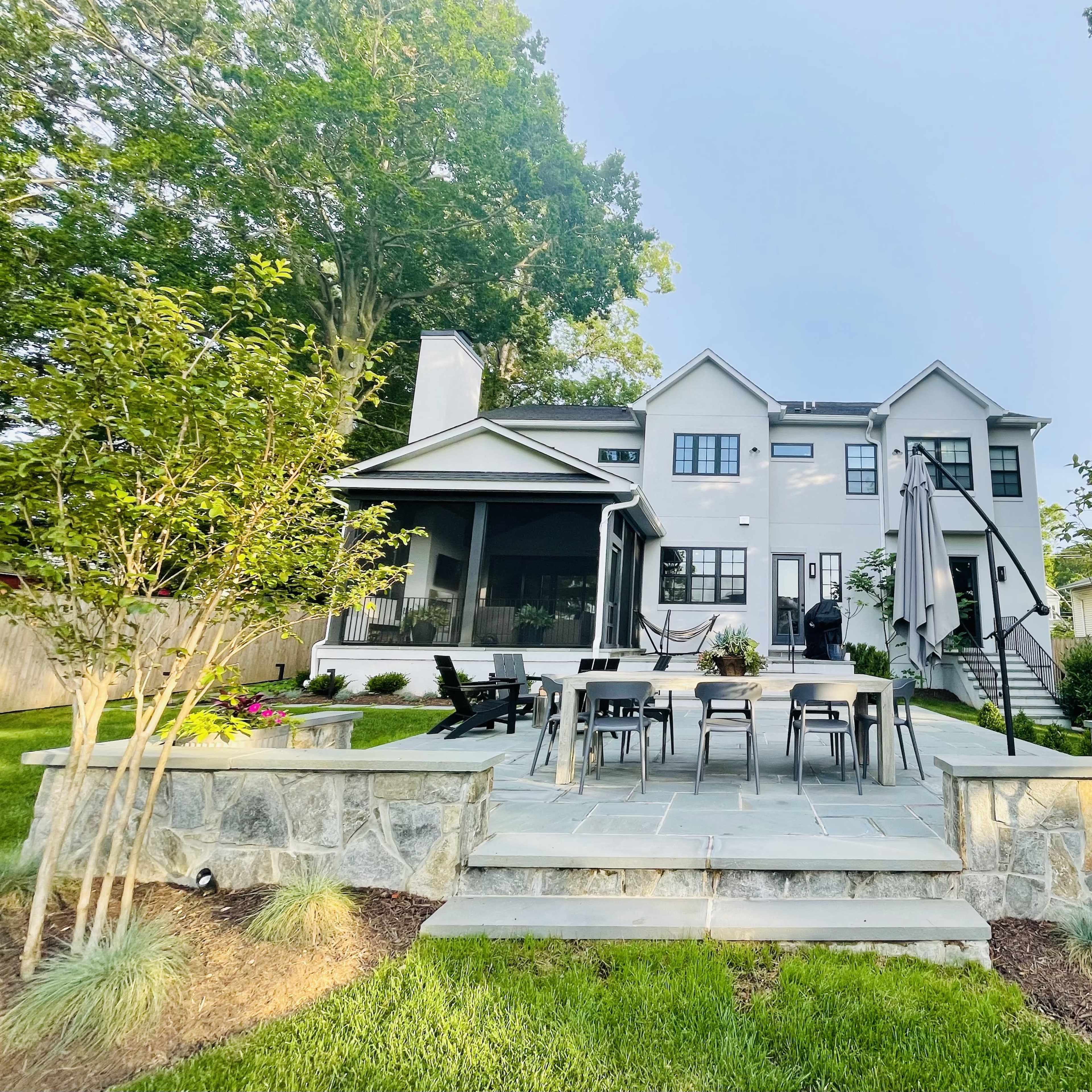 A spacious backyard features a stone patio with a dining table and chairs, leading up to a modern two-story home surrounded by greenery.