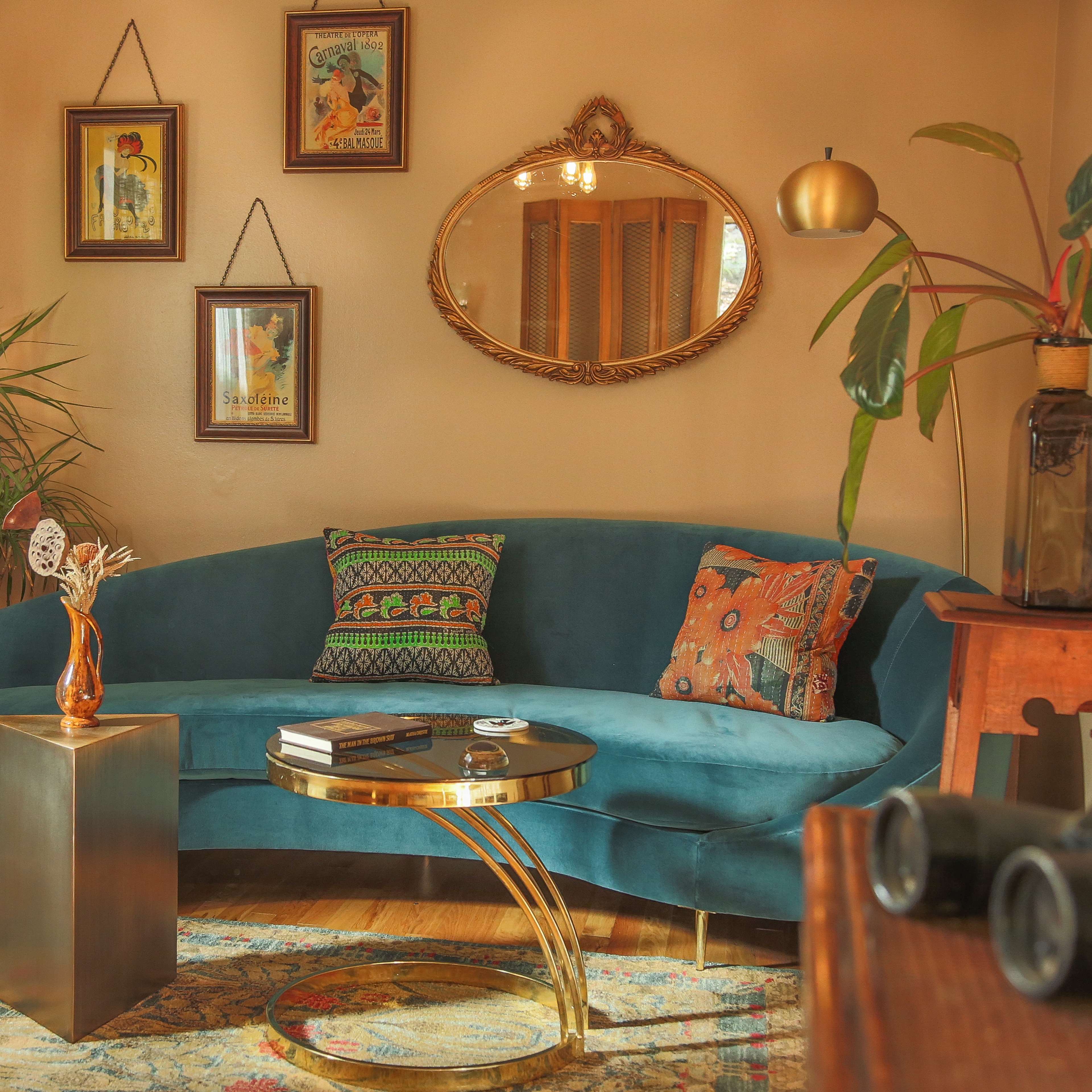 The scene features a curved teal sofa surrounded by framed artwork, a round coffee table, and decorative plants in a warmly lit living room.