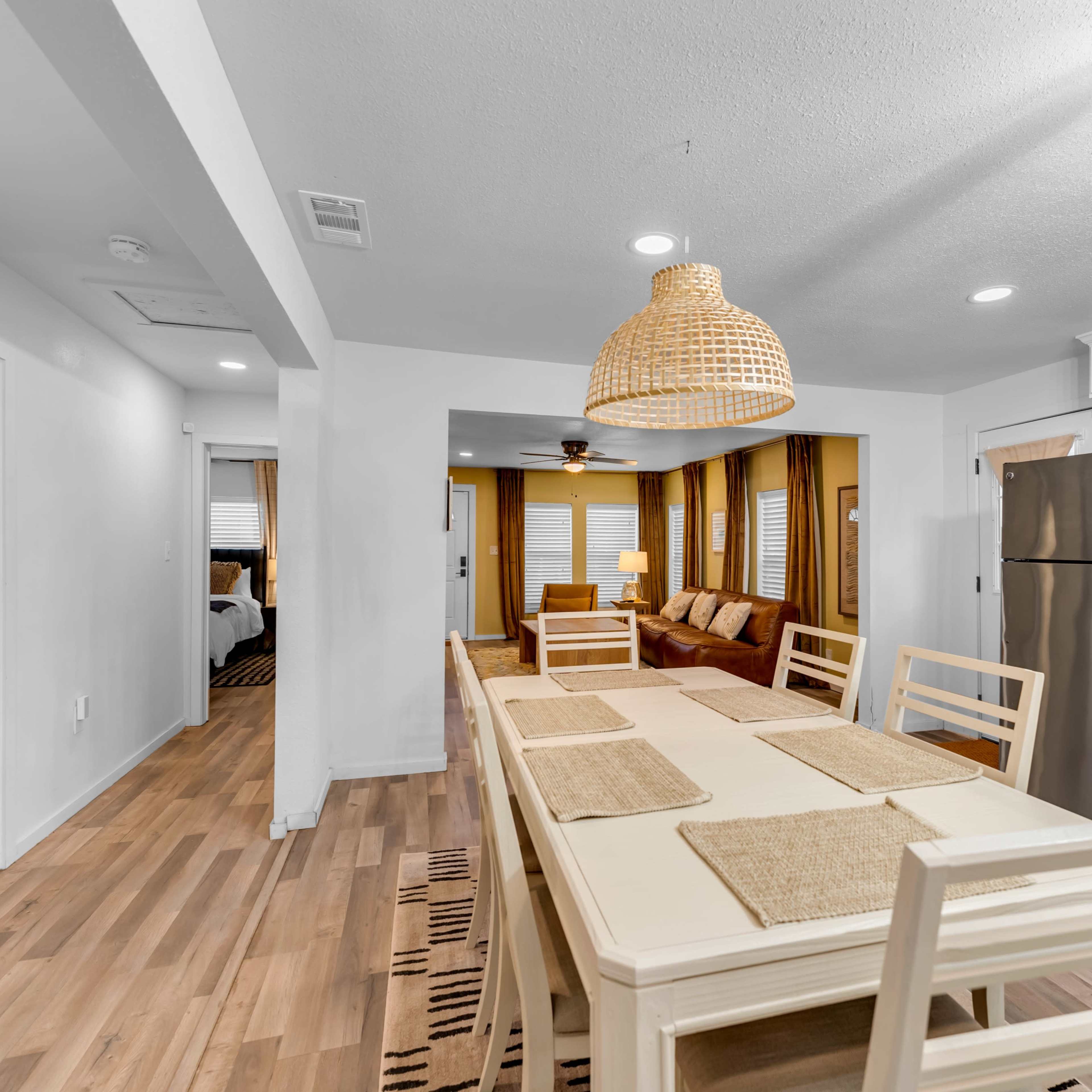 A modern dining area features a long wooden table with beige placemats, surrounded by chairs, with a kitchenette and living room visible in the background.