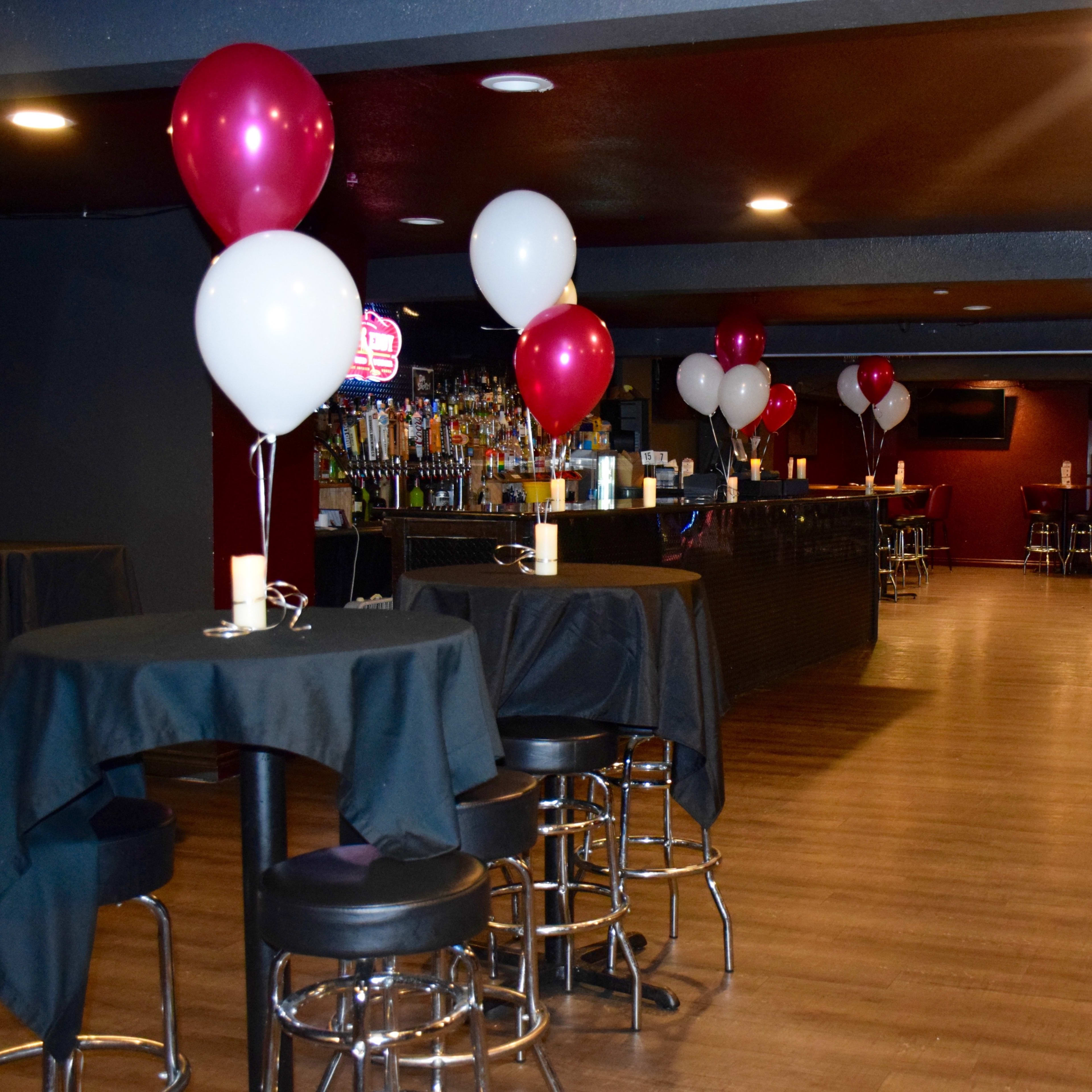 A dimly lit bar area with high-top tables covered in black cloths, featuring red and white balloons and a long bar lined with bottles in the background.