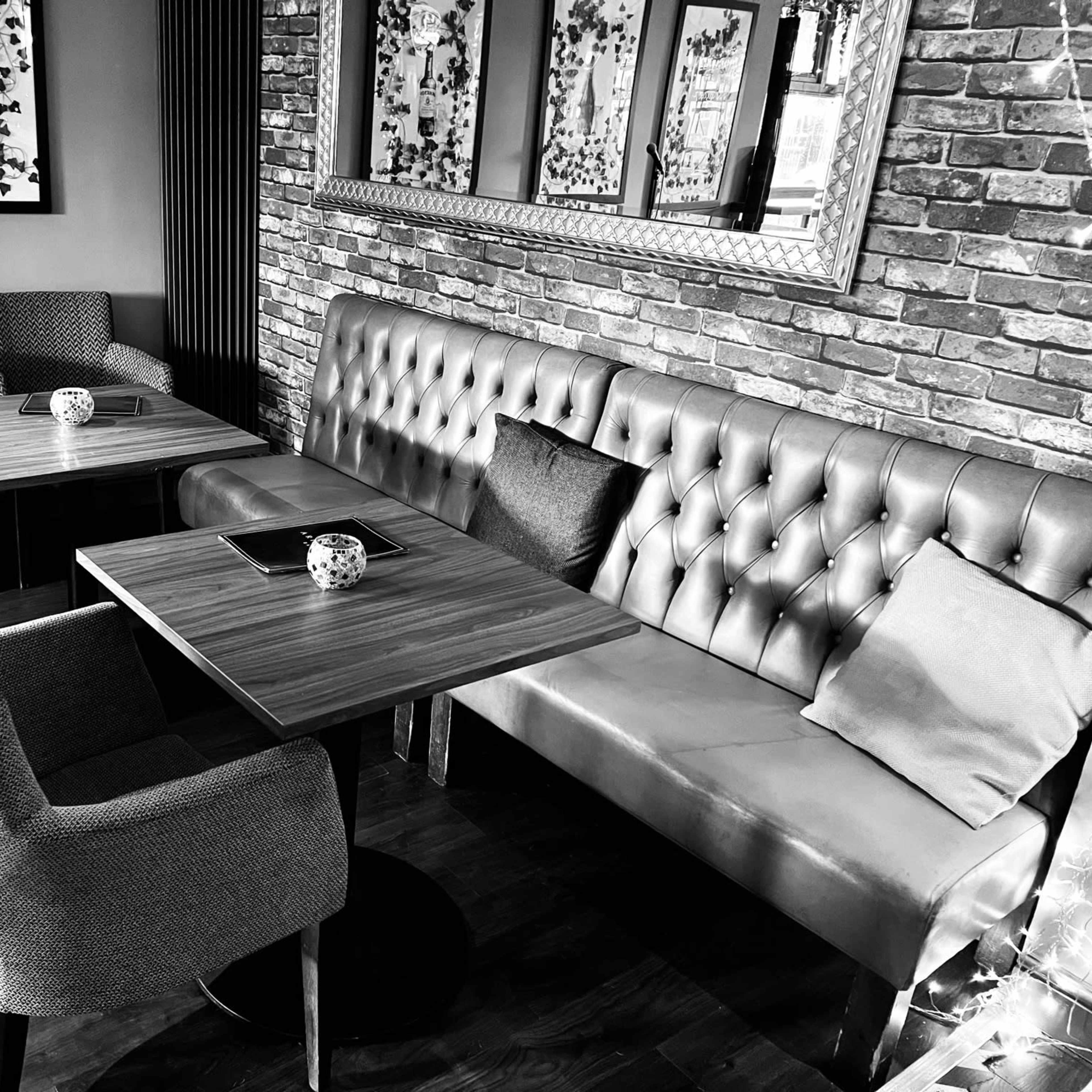A cozy seating area with two tables and a mix of upholstered chairs and a tufted bench against a brick wall, framed by decorative mirrors.