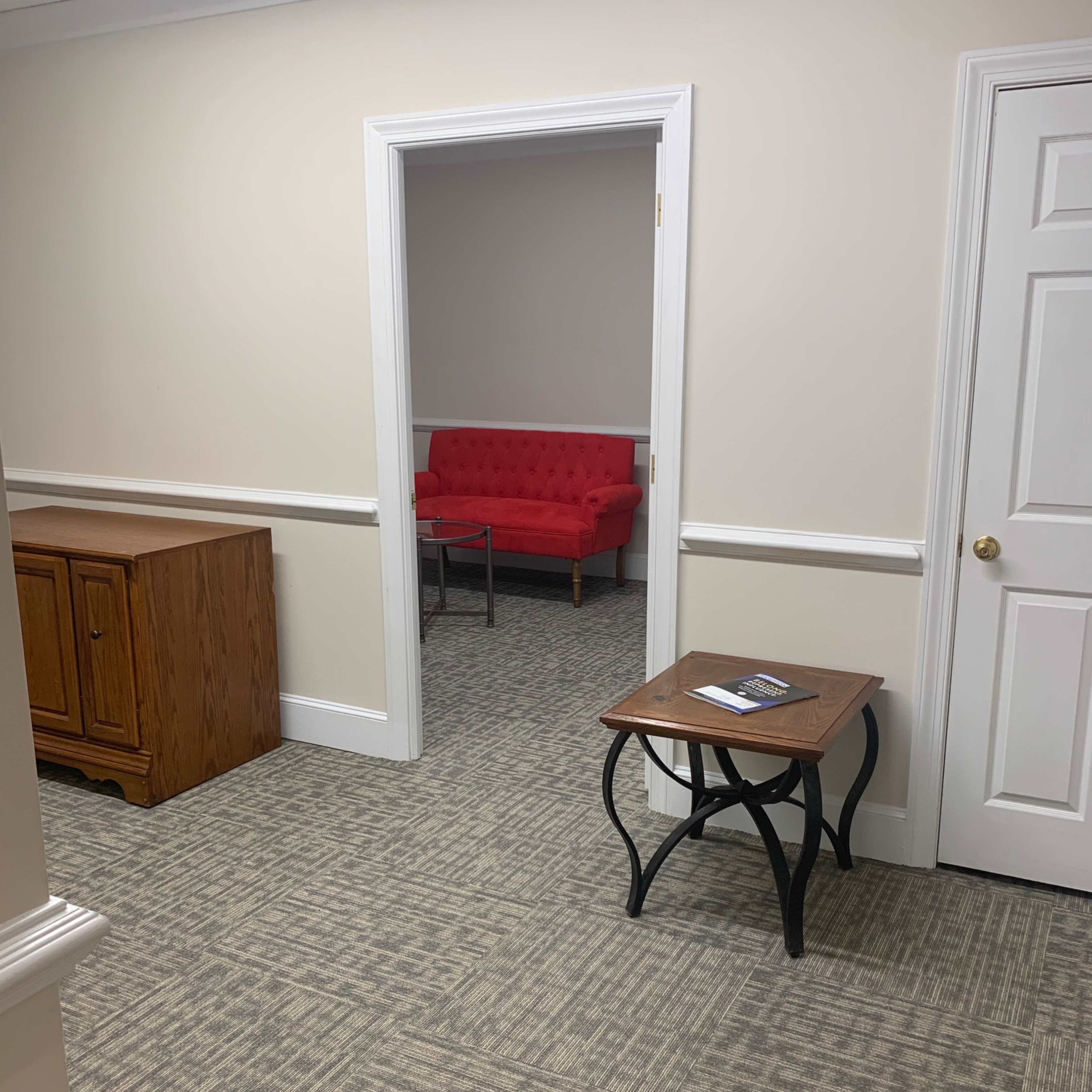 A hallway leading to a room with a red couch visible in the background, next to a wooden cabinet and a small table in the foreground.