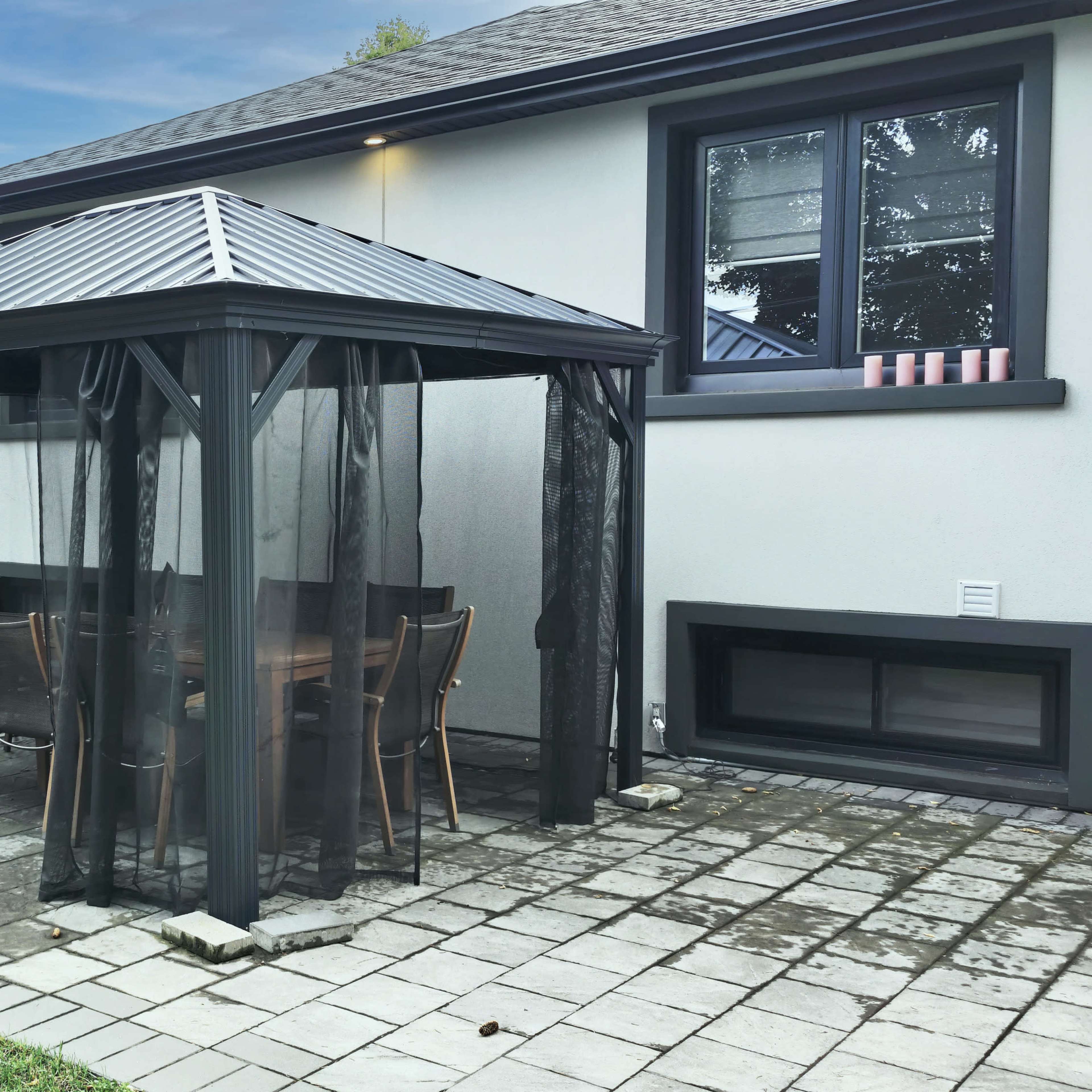 A black gazebo with mesh curtains is situated on a paved patio, alongside a hammock and a wall with a window.