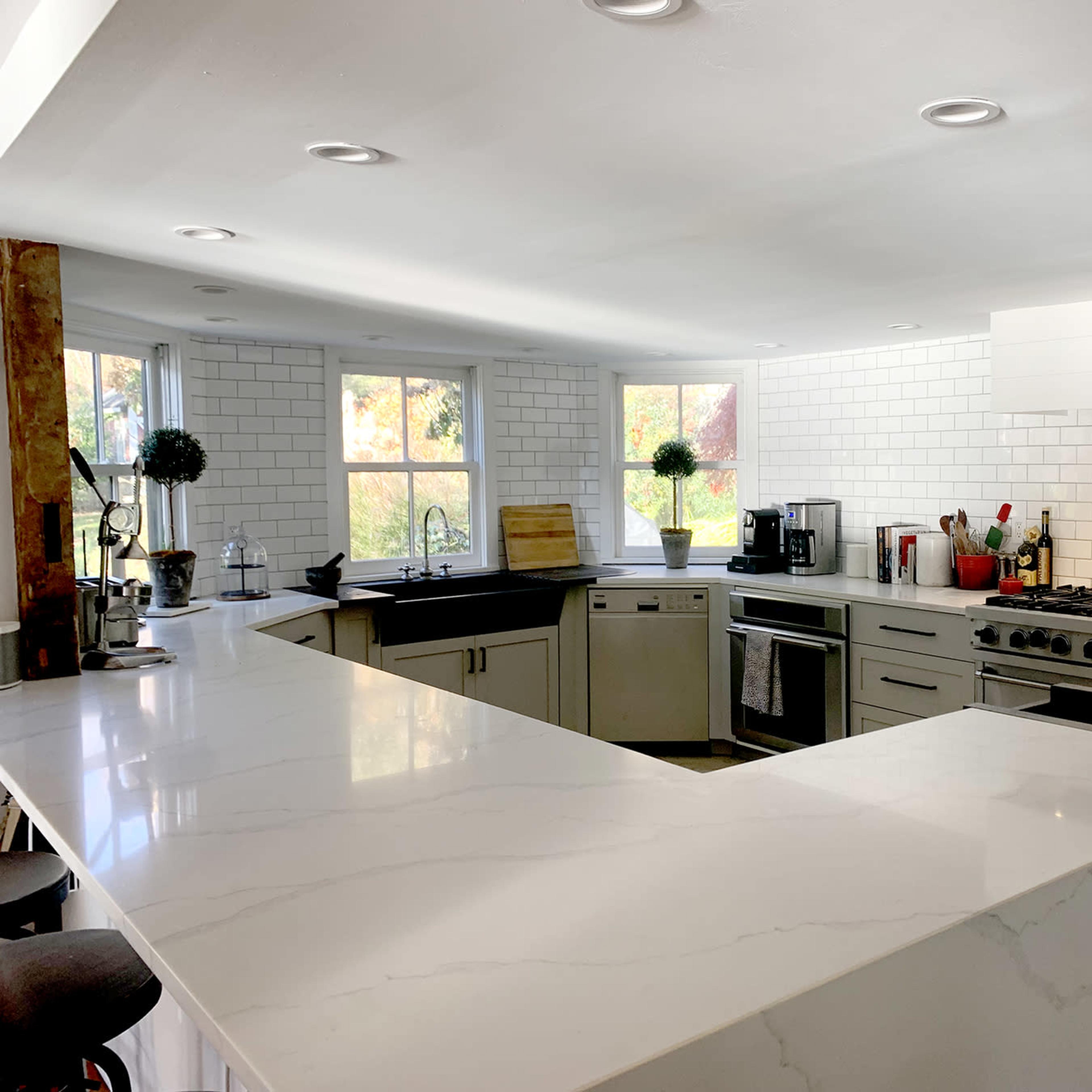 A modern kitchen with a large white island, stainless steel appliances, and white tile backsplash, featuring a bright atmosphere from multiple windows.