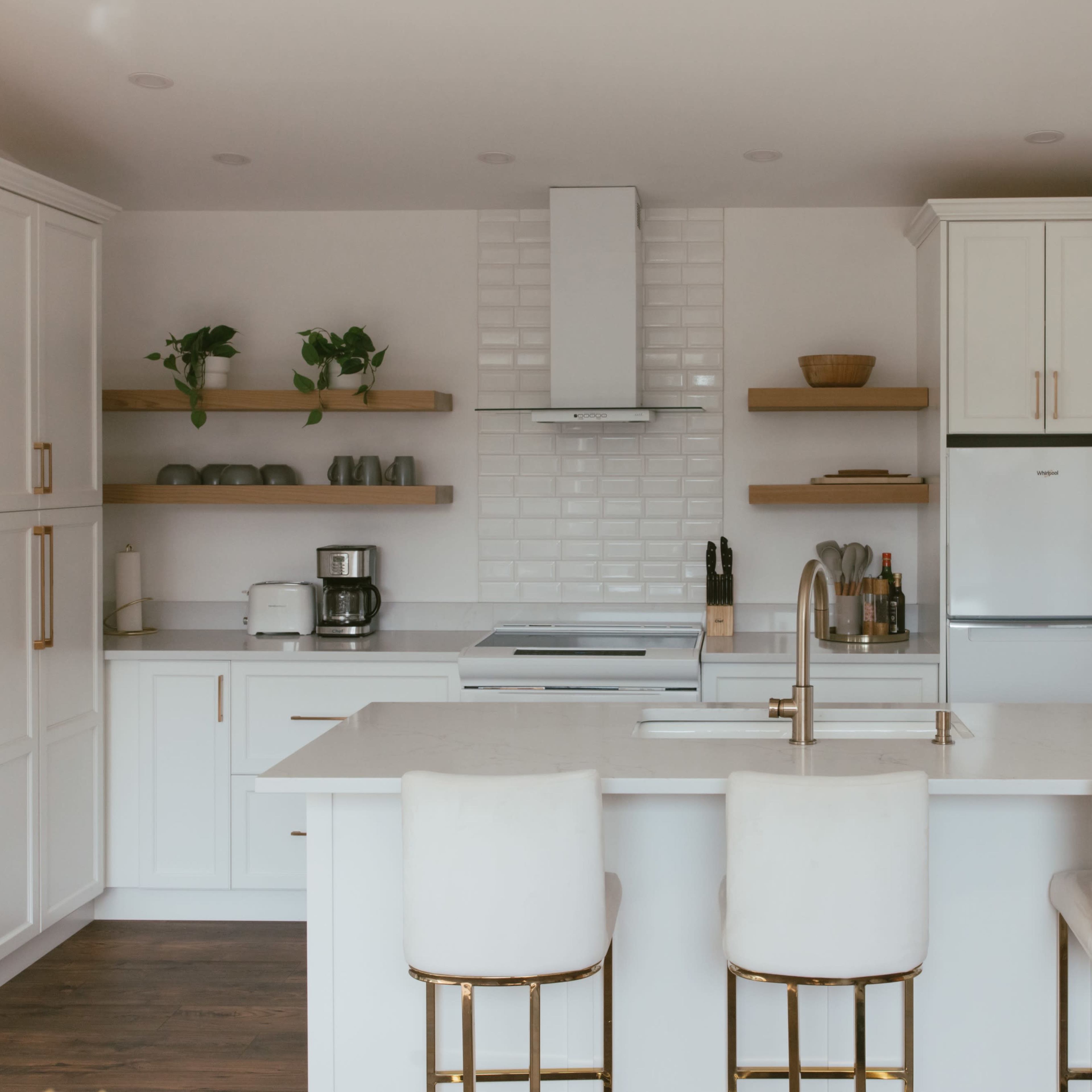A modern kitchen features white cabinetry, a large island with bar stools, and open shelving with plants and kitchenware.