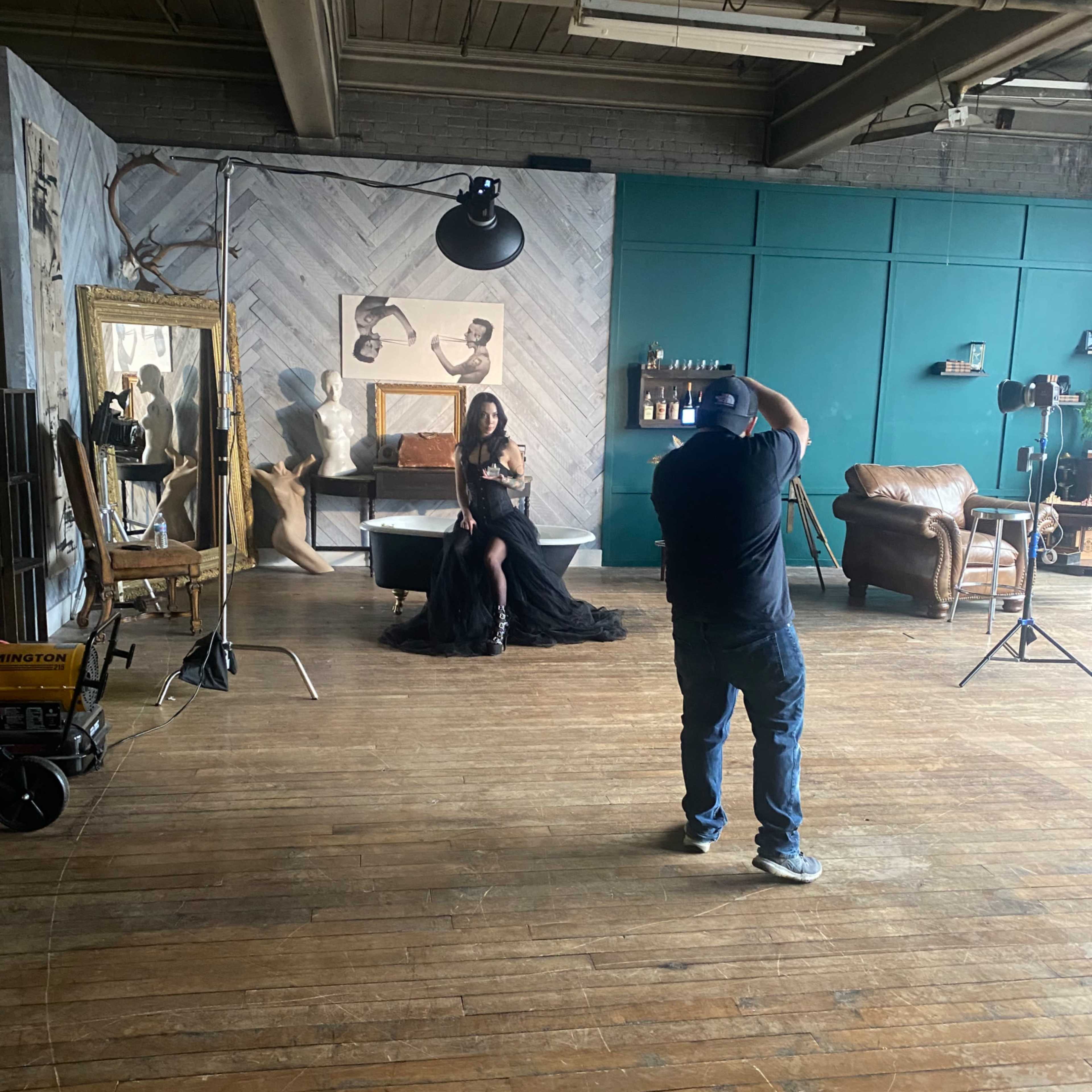 A photographer captures an image of a woman in a black dress while she poses in a well-lit studio with wooden floors and studio equipment.