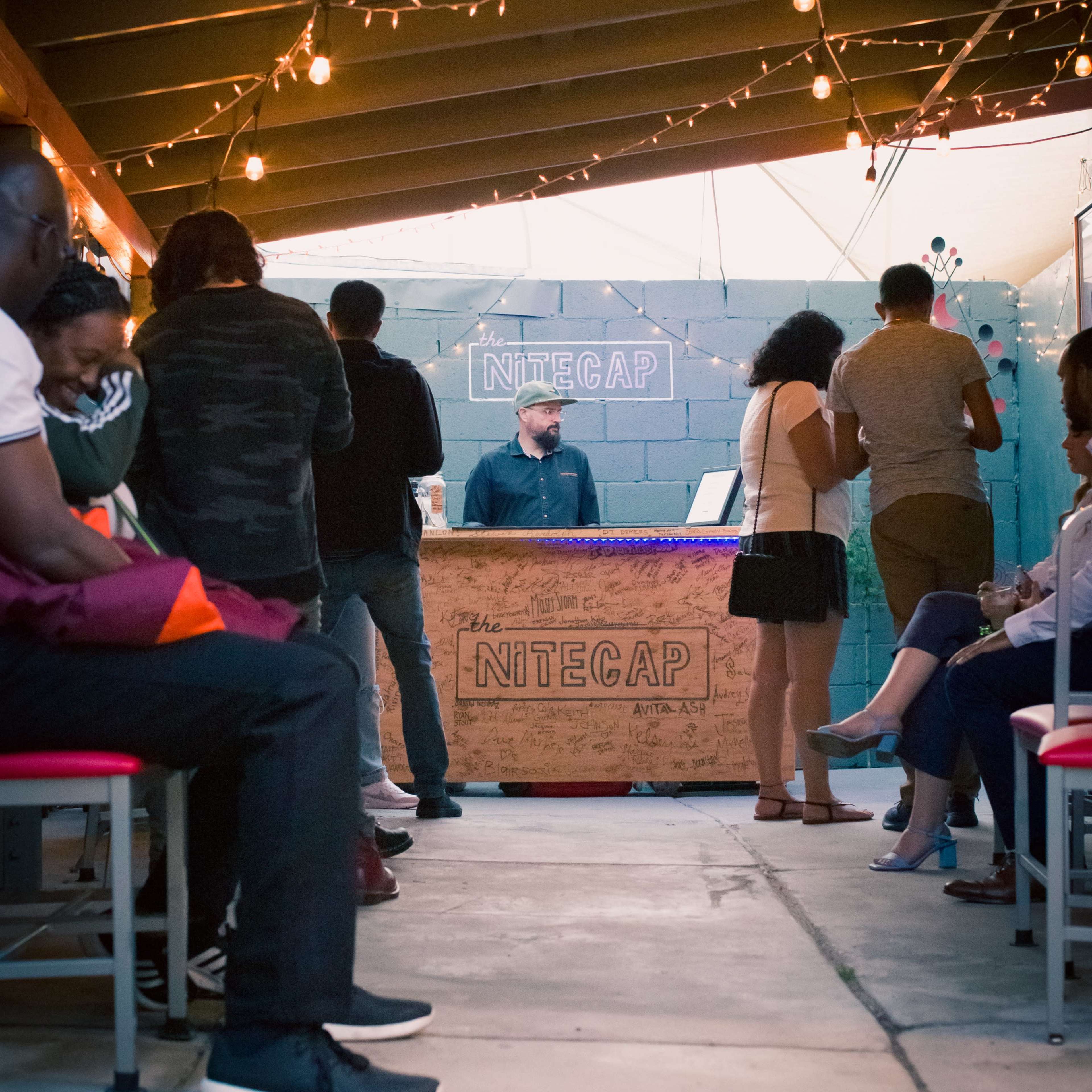 A dimly lit outdoor bar scene features patrons seated on benches while a bartender serves drinks at a counter labeled "NiteCap."