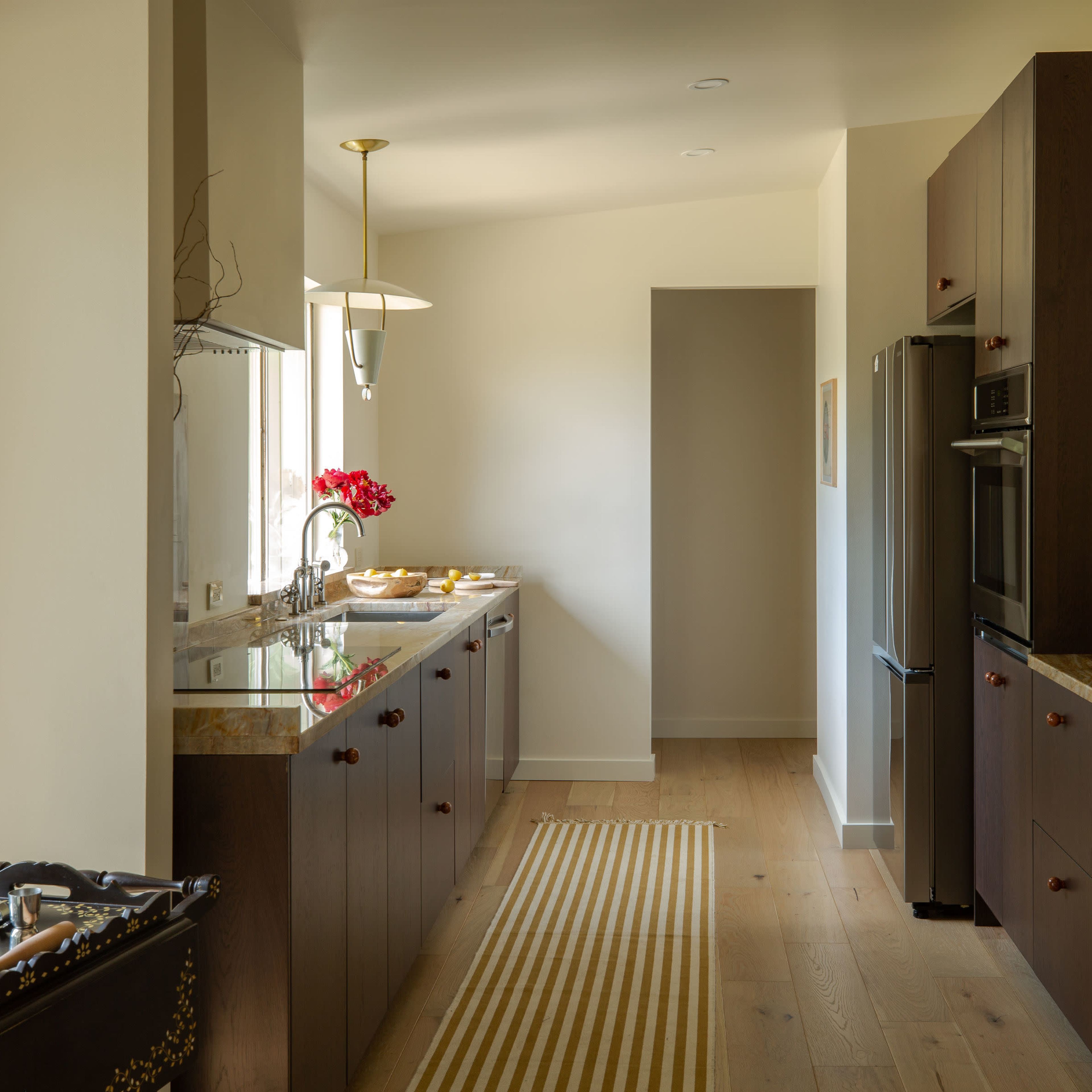 A modern kitchen features dark wood cabinets, a row of recessed lights, and a striped rug running along the polished wooden floor.