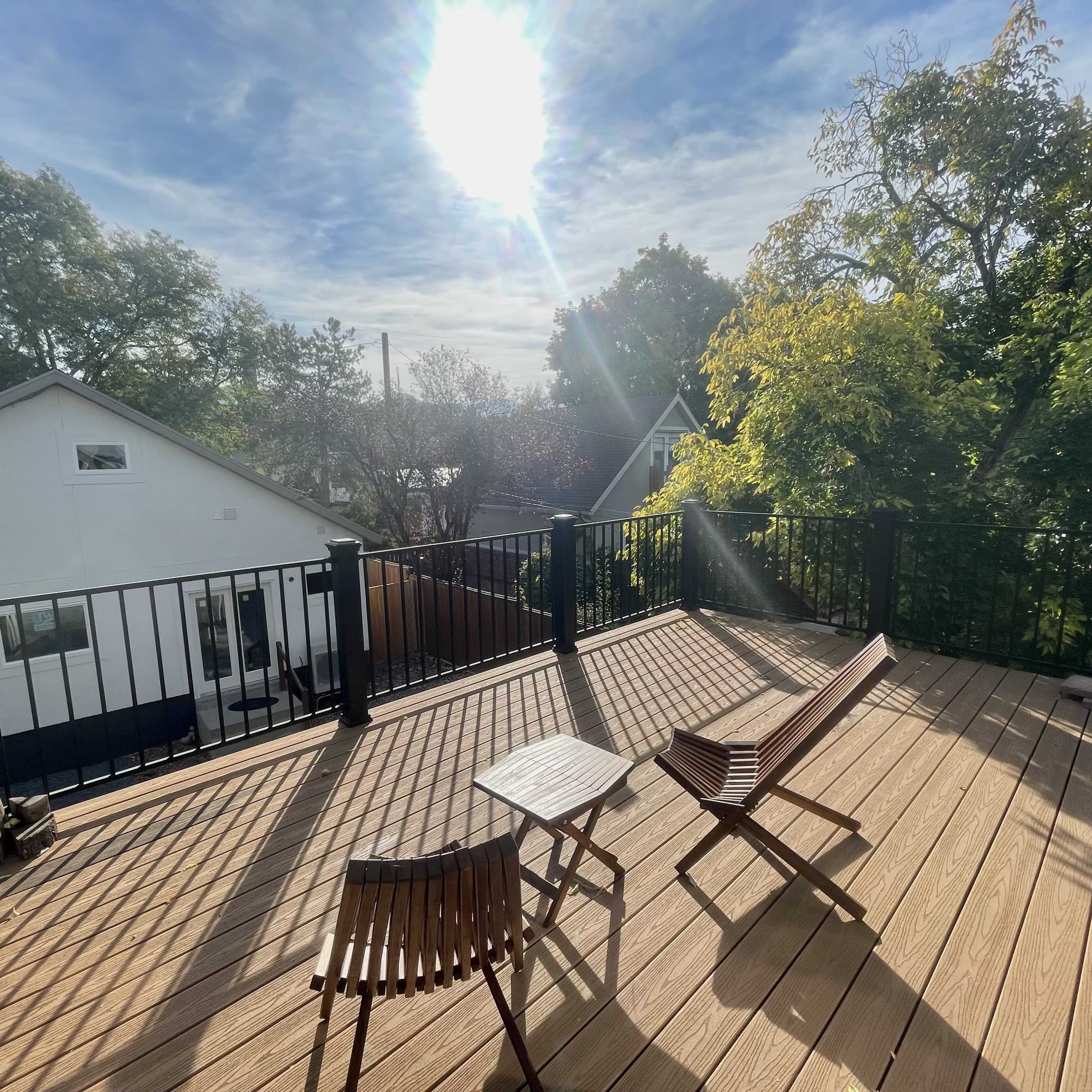A sunny deck with two wooden chairs and a small table, overlooking green trees and a house in the background.