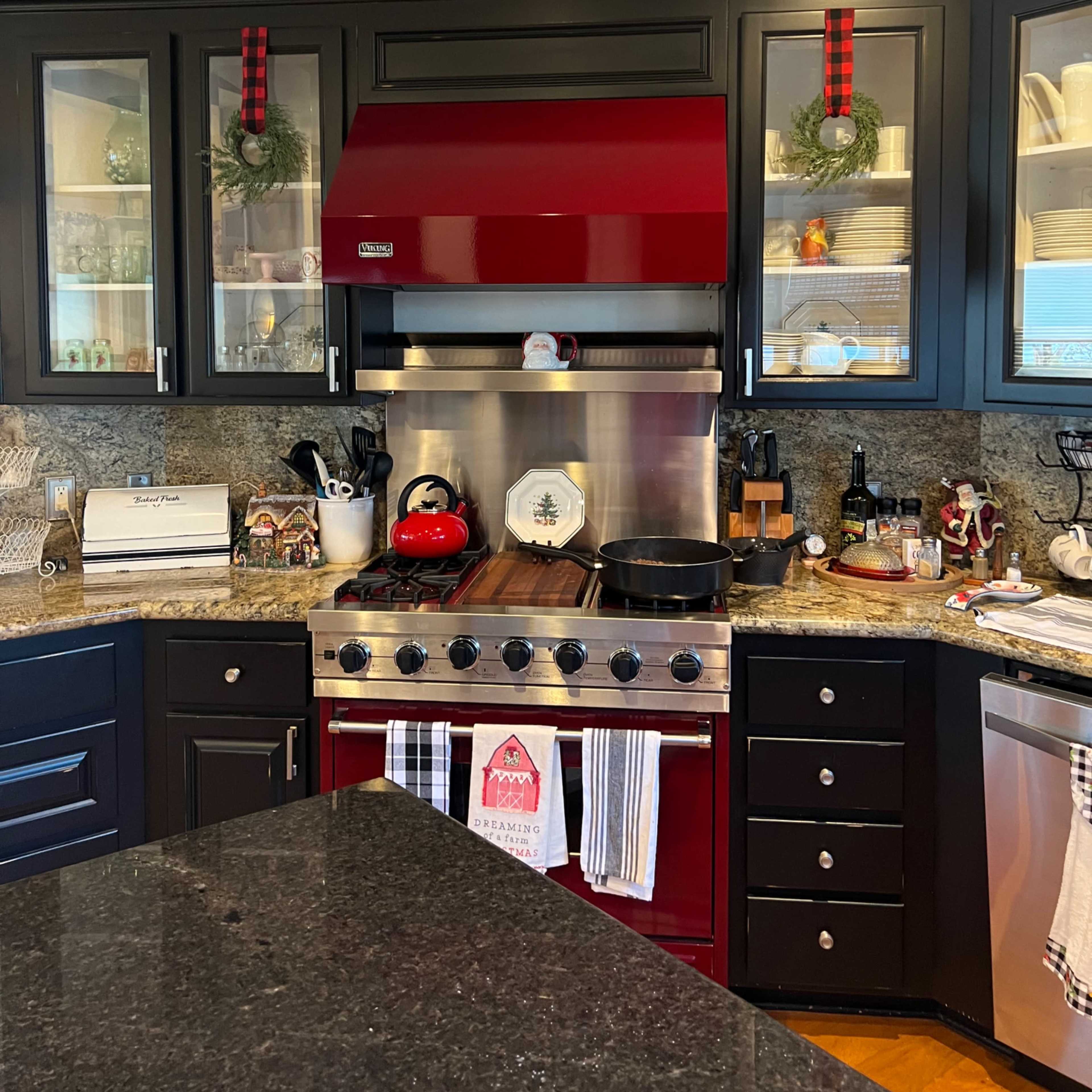 The kitchen features a red range hood above a stainless steel stove, surrounded by dark cabinets, open shelving with dishware, and decorations for the holiday season.