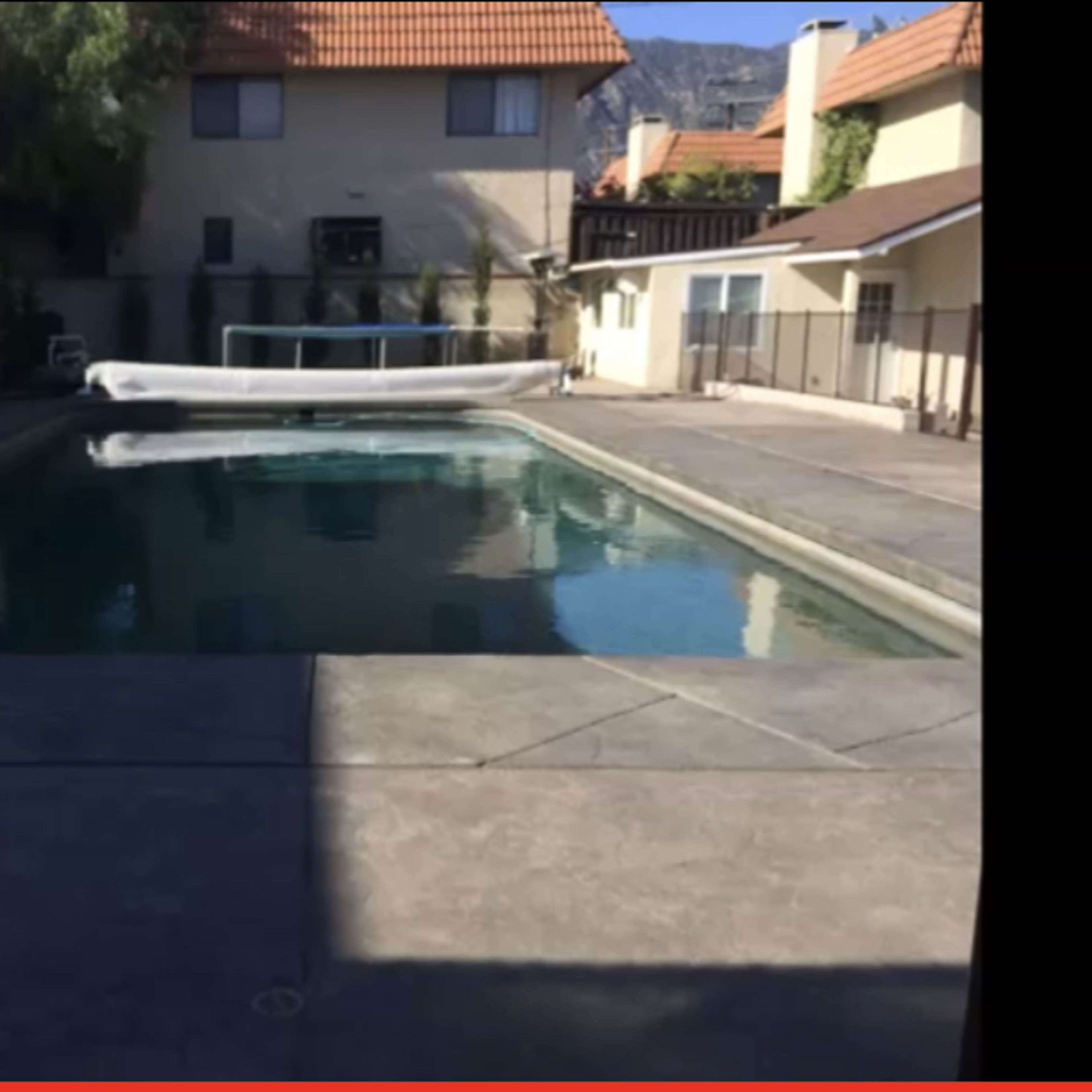 A rectangular swimming pool surrounded by a concrete deck and residential buildings with a sloped roof in the background.