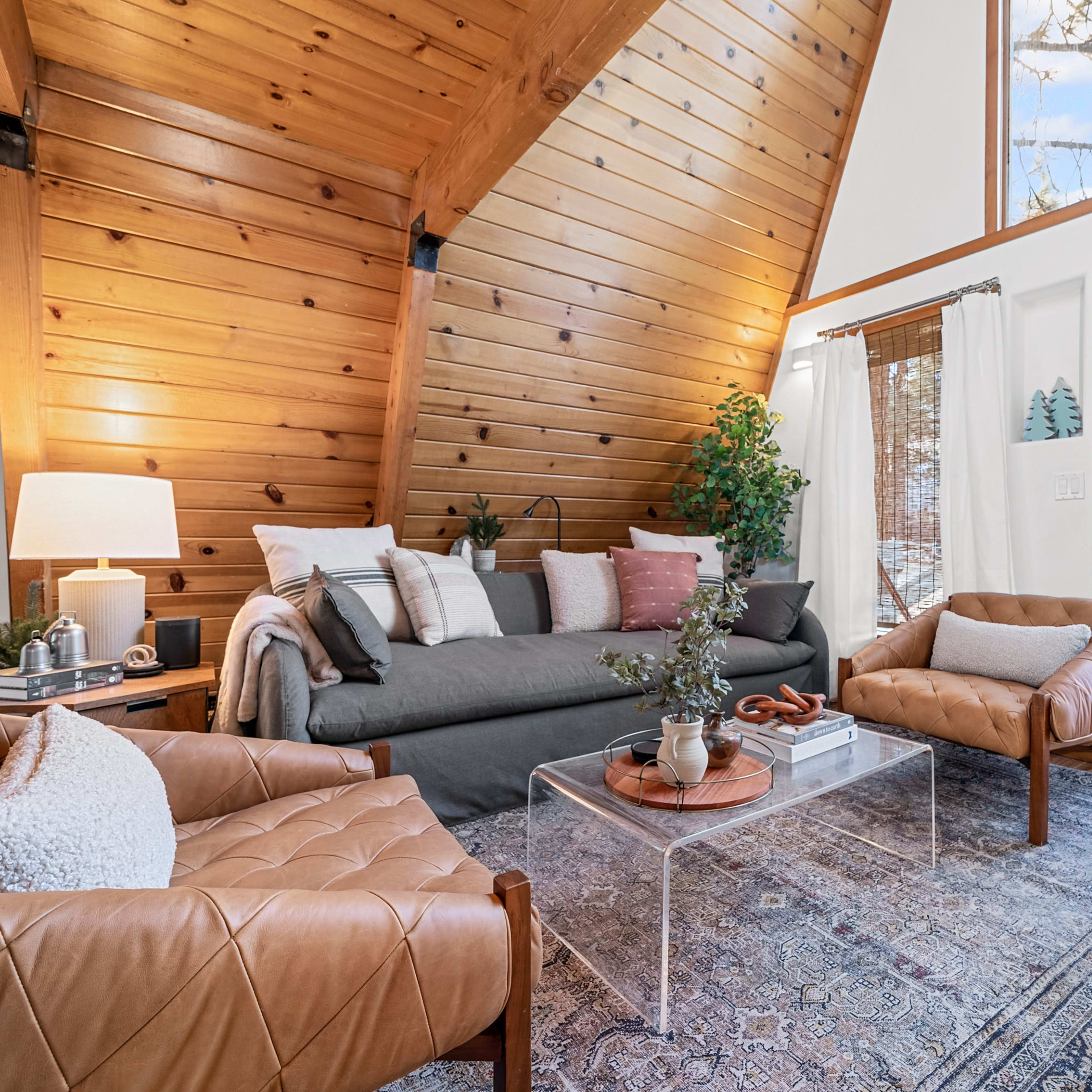 A cozy living room with a high wooden ceiling, a grey sofa, leather chairs, and a glass coffee table on a patterned rug, complemented by a door that opens to the outside.