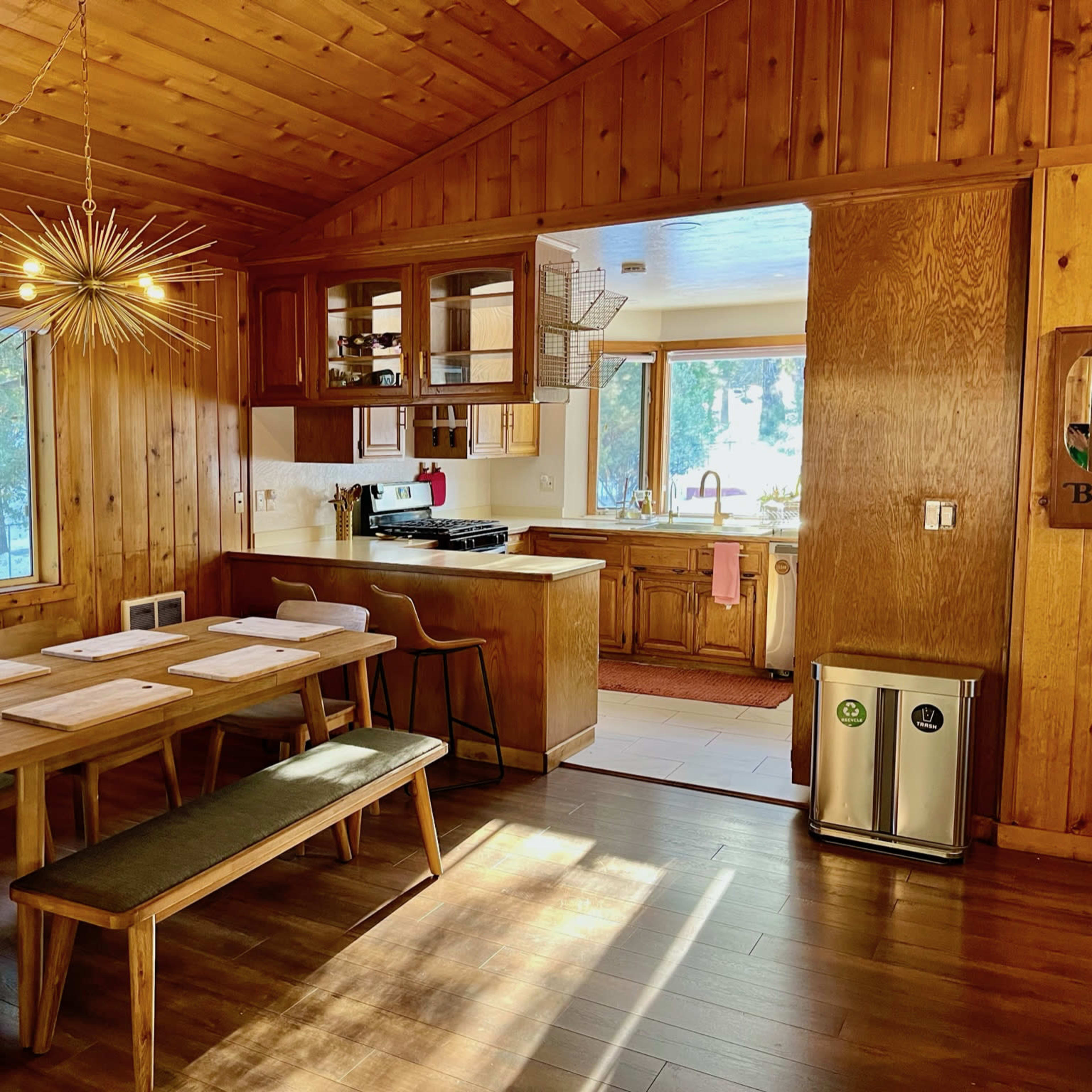 A wooden-paneled dining area with a table, benches, and a kitchen visible in the background.