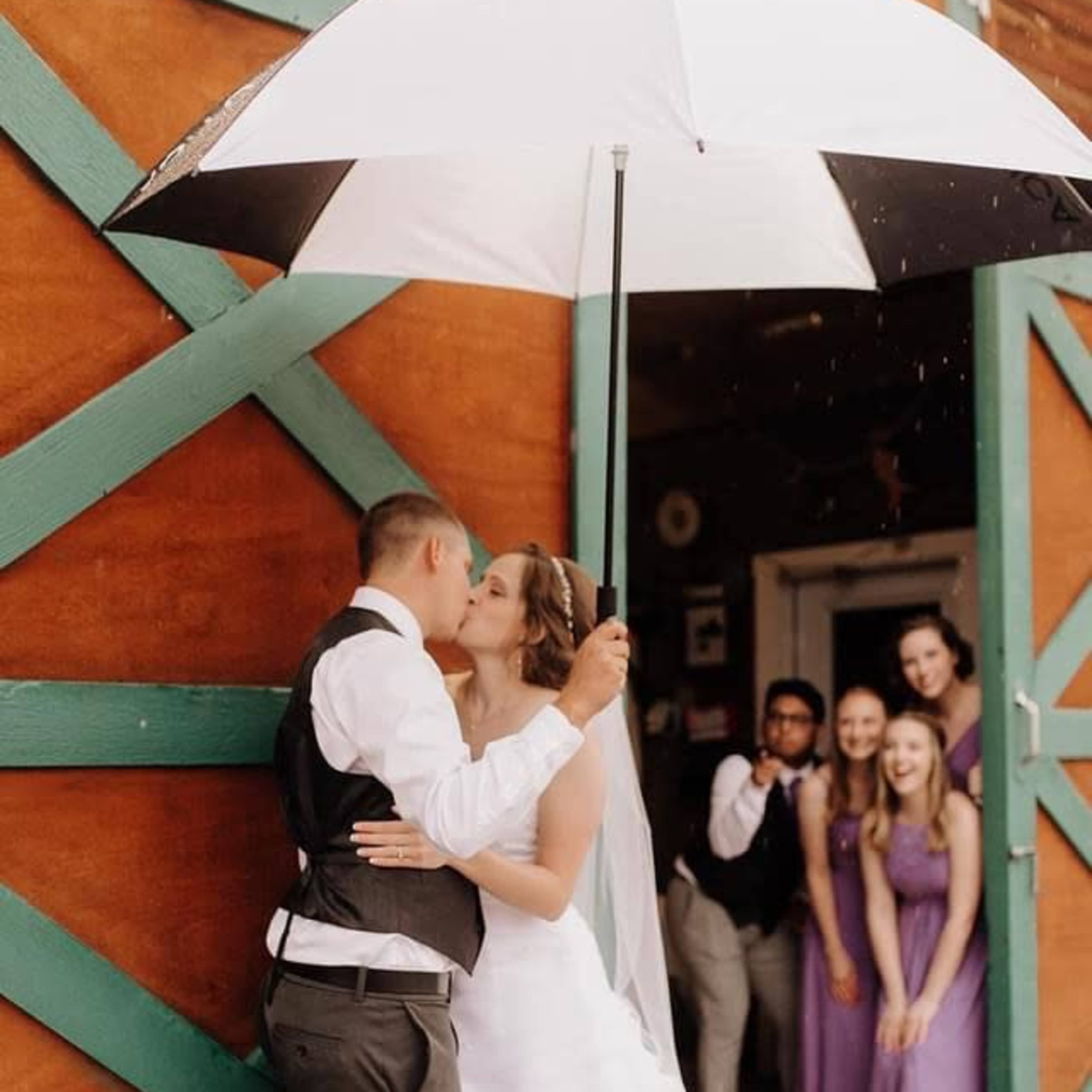 A couple in wedding attire shares a kiss under a large umbrella, with a group of people in formal dress smiling in the background.