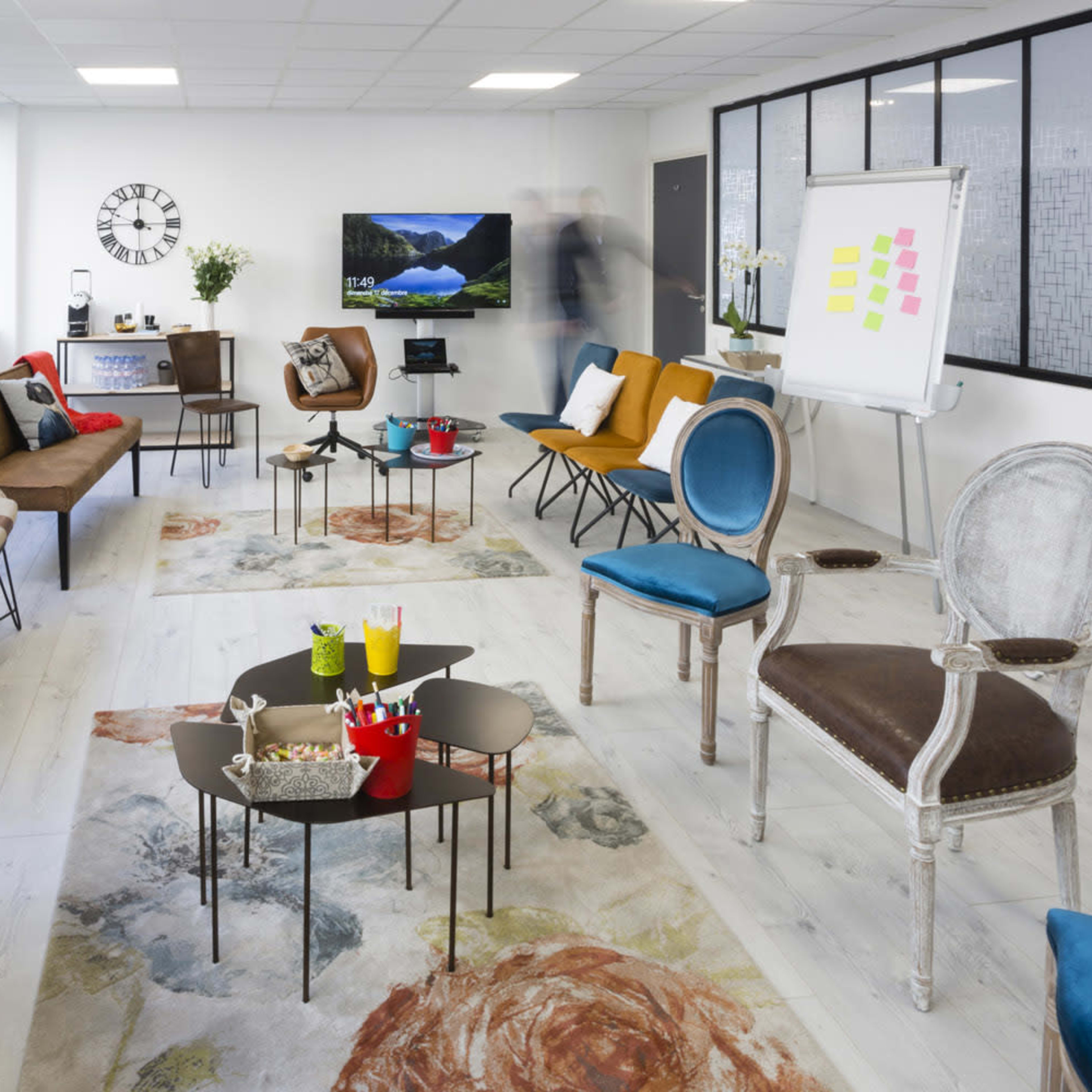 A modern, spacious meeting room filled with various seating options and tables, featuring large windows and a presentation area with a whiteboard.