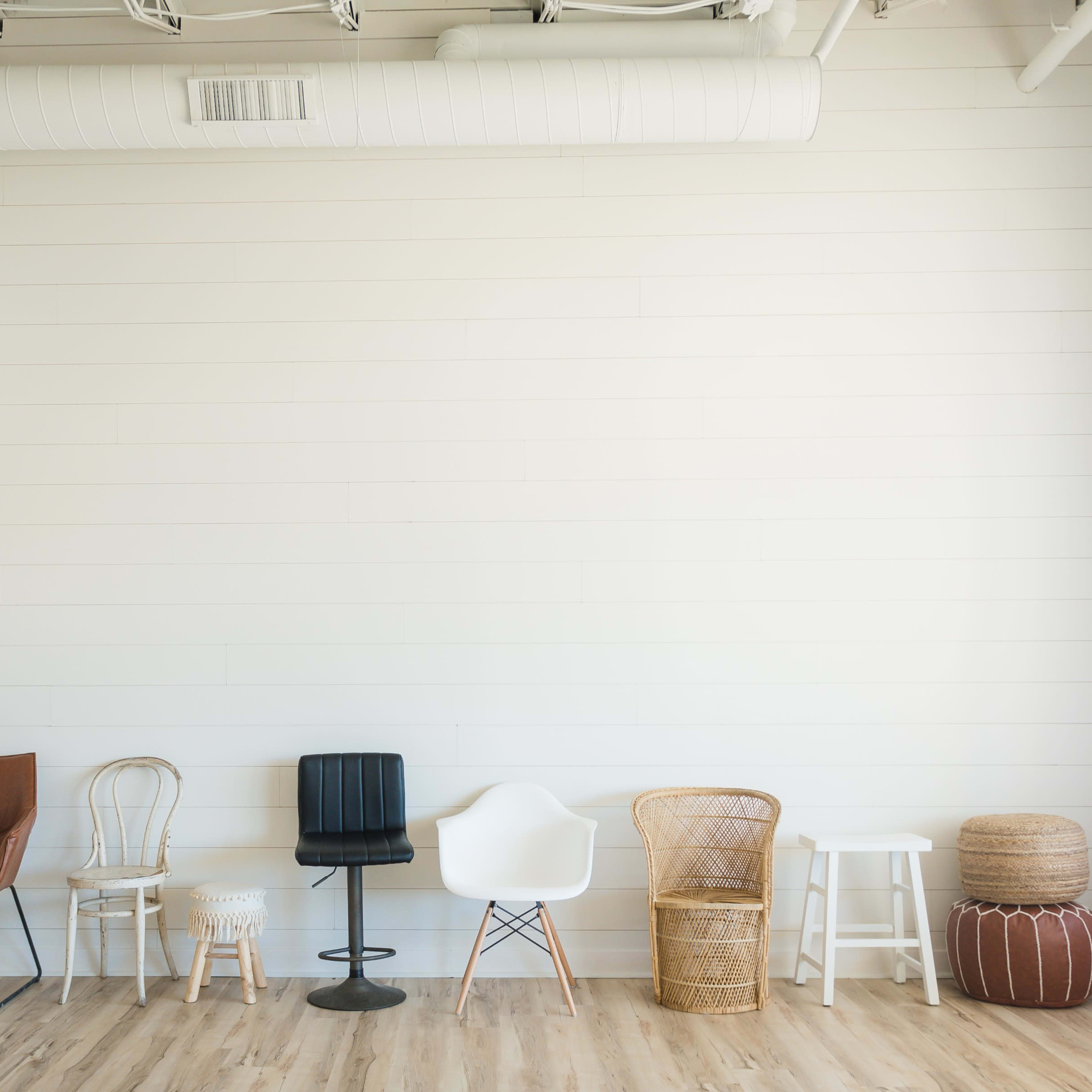 A row of five different styles of chairs and stools against a plain, white wall in a well-lit room.