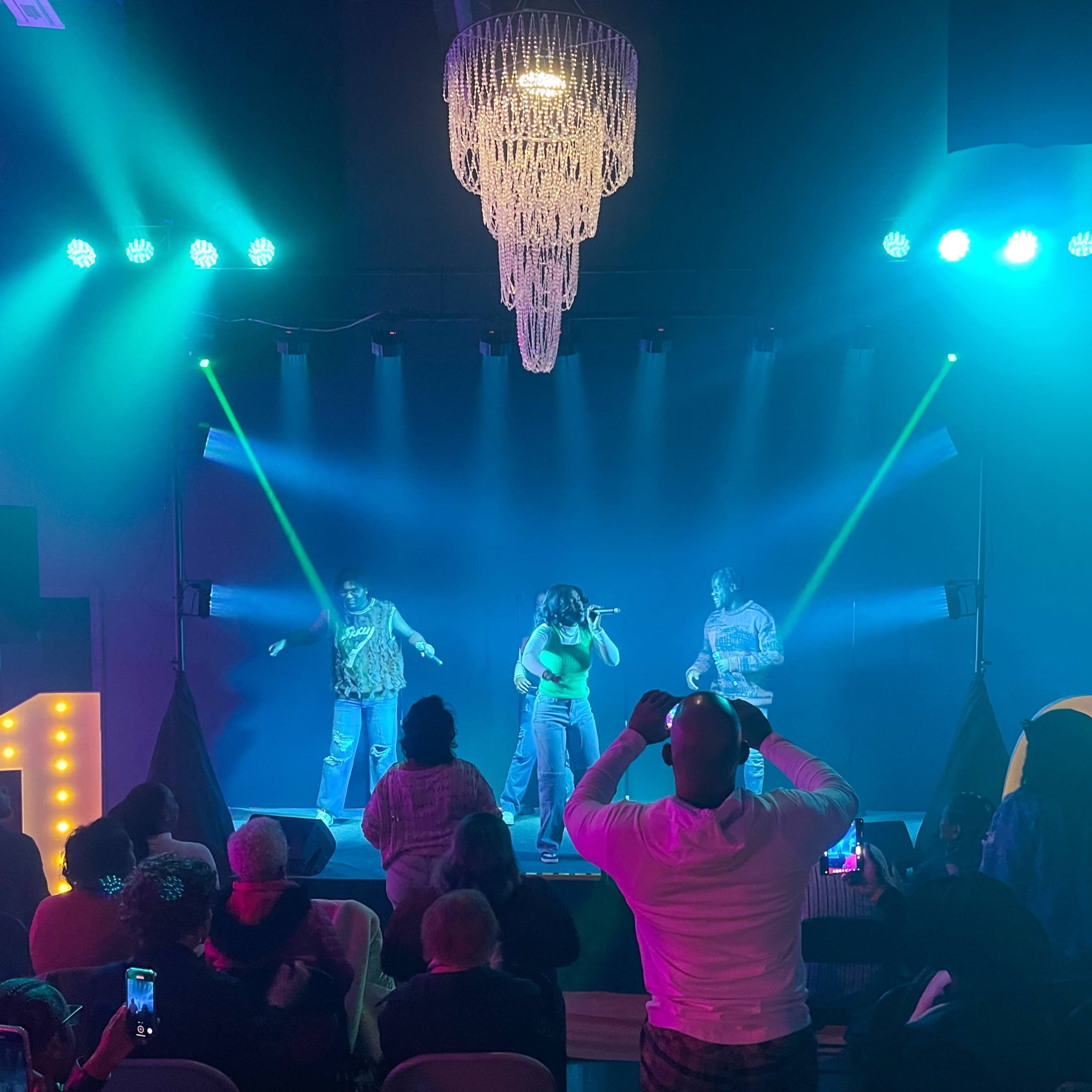 A live performance takes place on stage, illuminated by colorful lights, with an audience seated in front.
