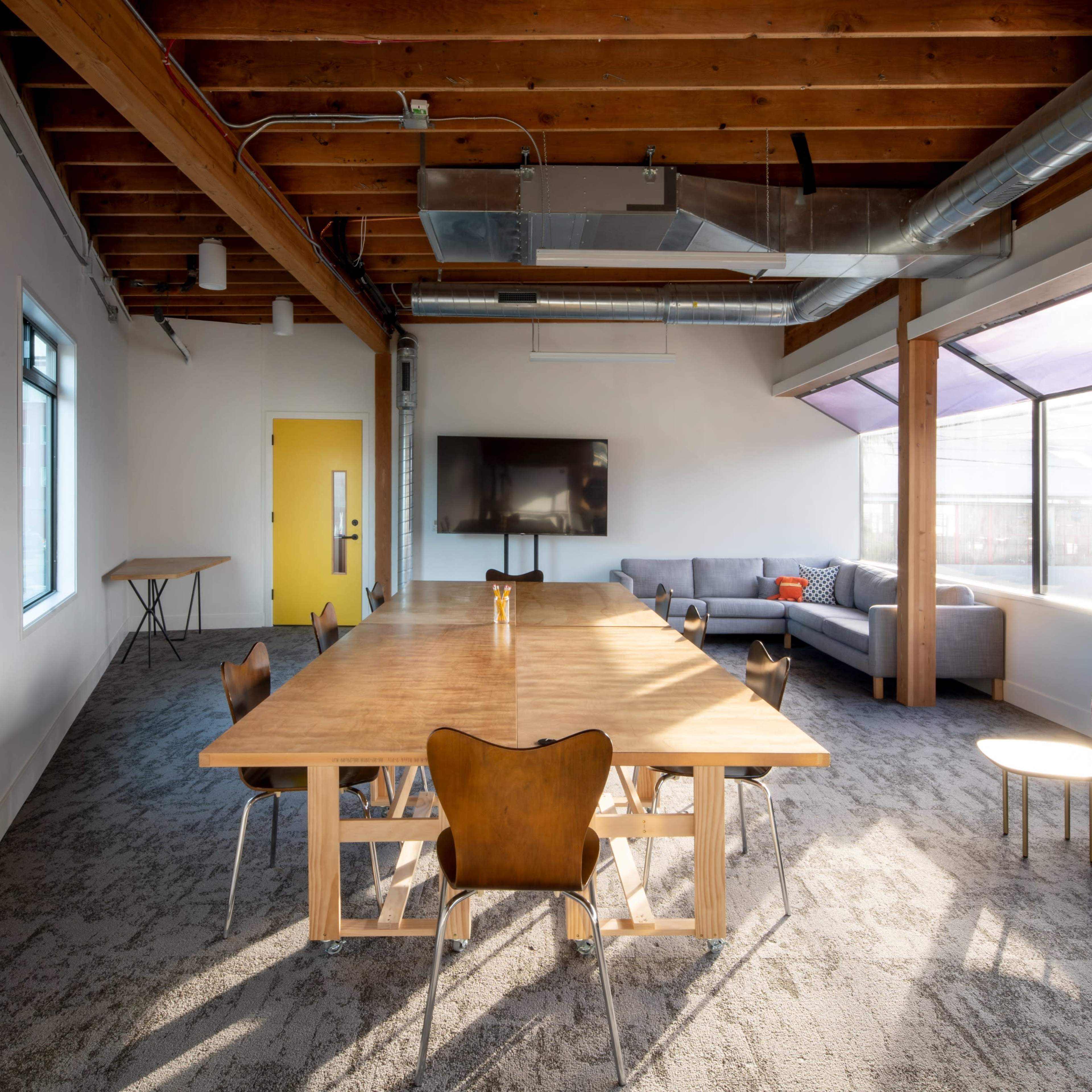 A spacious conference room features a large wooden table surrounded by chairs, with a seating area and large windows allowing natural light.