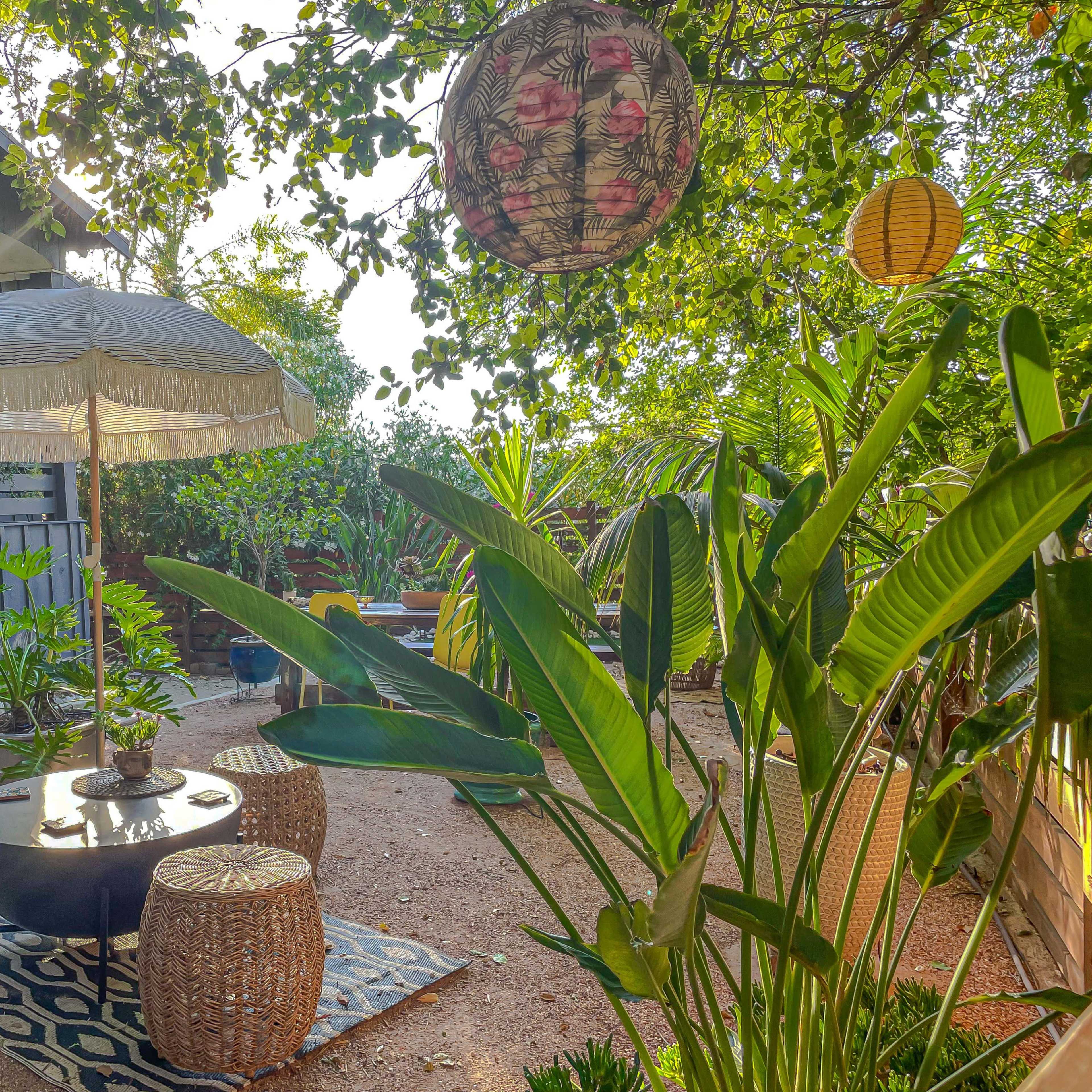 A shaded outdoor seating area features a table surrounded by decorative stools, with hanging lanterns overhead amidst green plants.