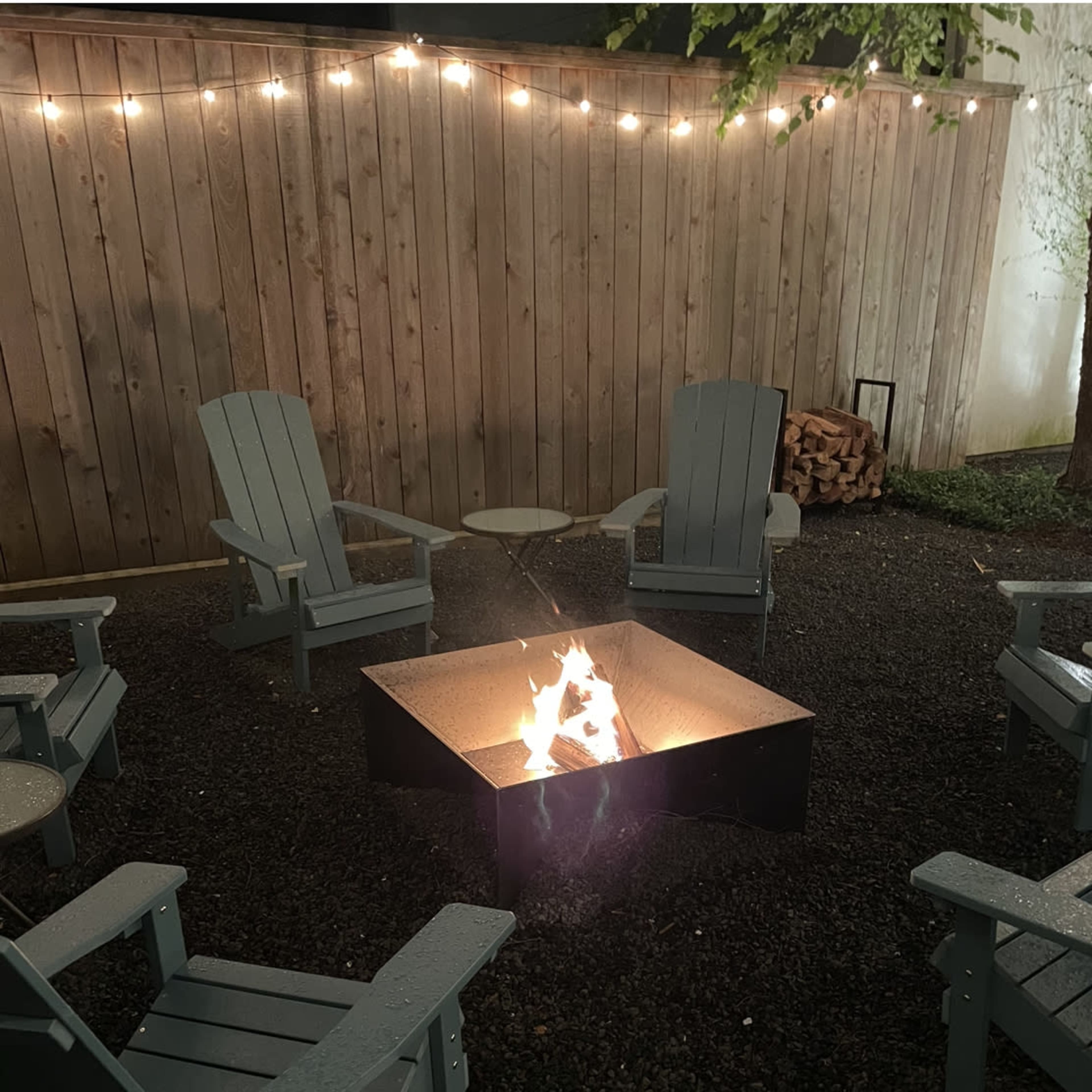 A fire pit surrounded by six adirondack chairs is illuminated by string lights hanging above.