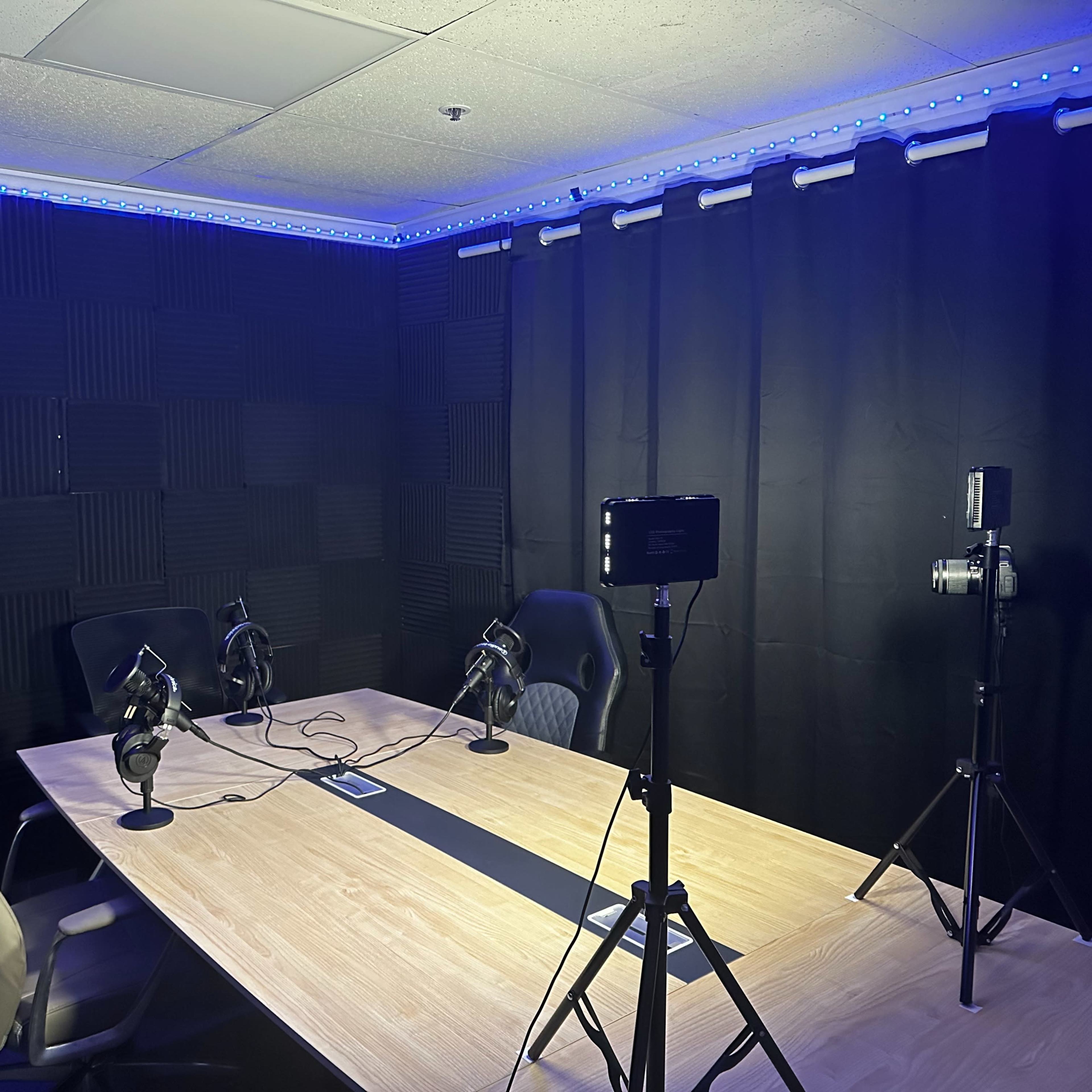 A recording studio featuring a large wooden table with several microphones, chairs, and lighting equipment, surrounded by sound-absorbing wall panels.
