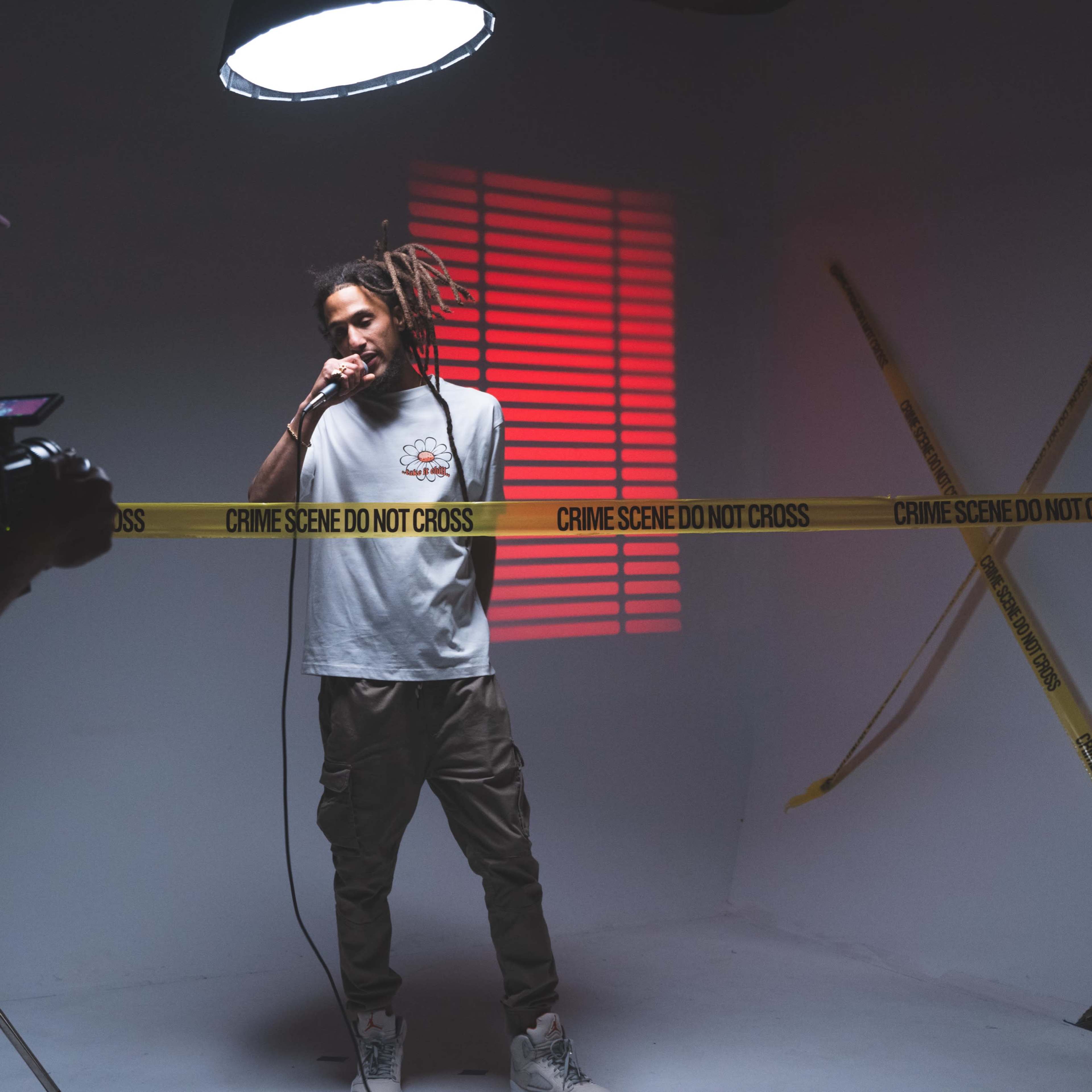 A male artist performs with a microphone in front of a camera, surrounded by yellow caution tape marked "CRIME SCENE DO NOT CROSS" and illuminated by a spotlight.