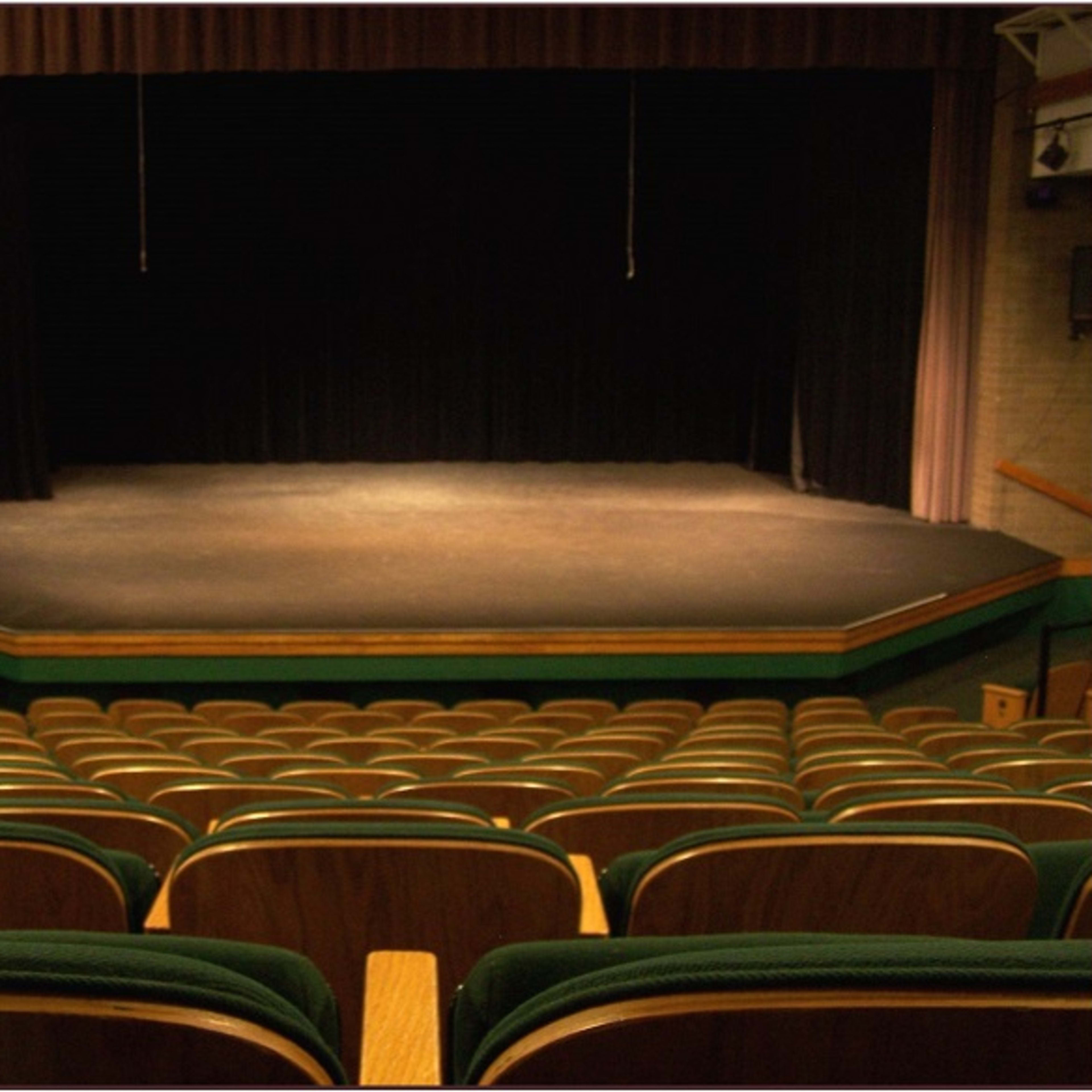 A darkened theater stage with a simple black curtain and rows of empty wooden seats in the foreground.