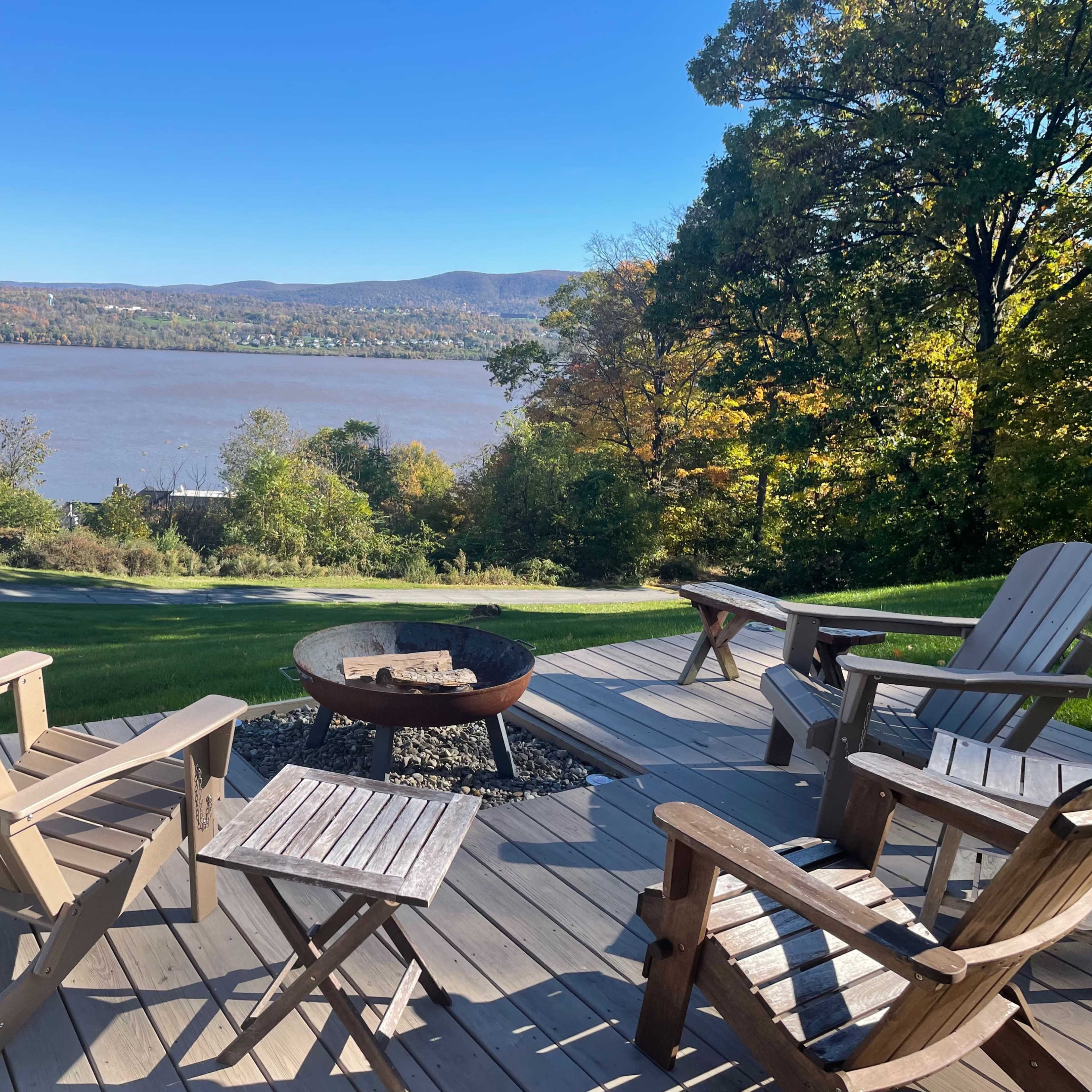 A seating area with wooden chairs and a fire pit overlooks a river and hills in the background.