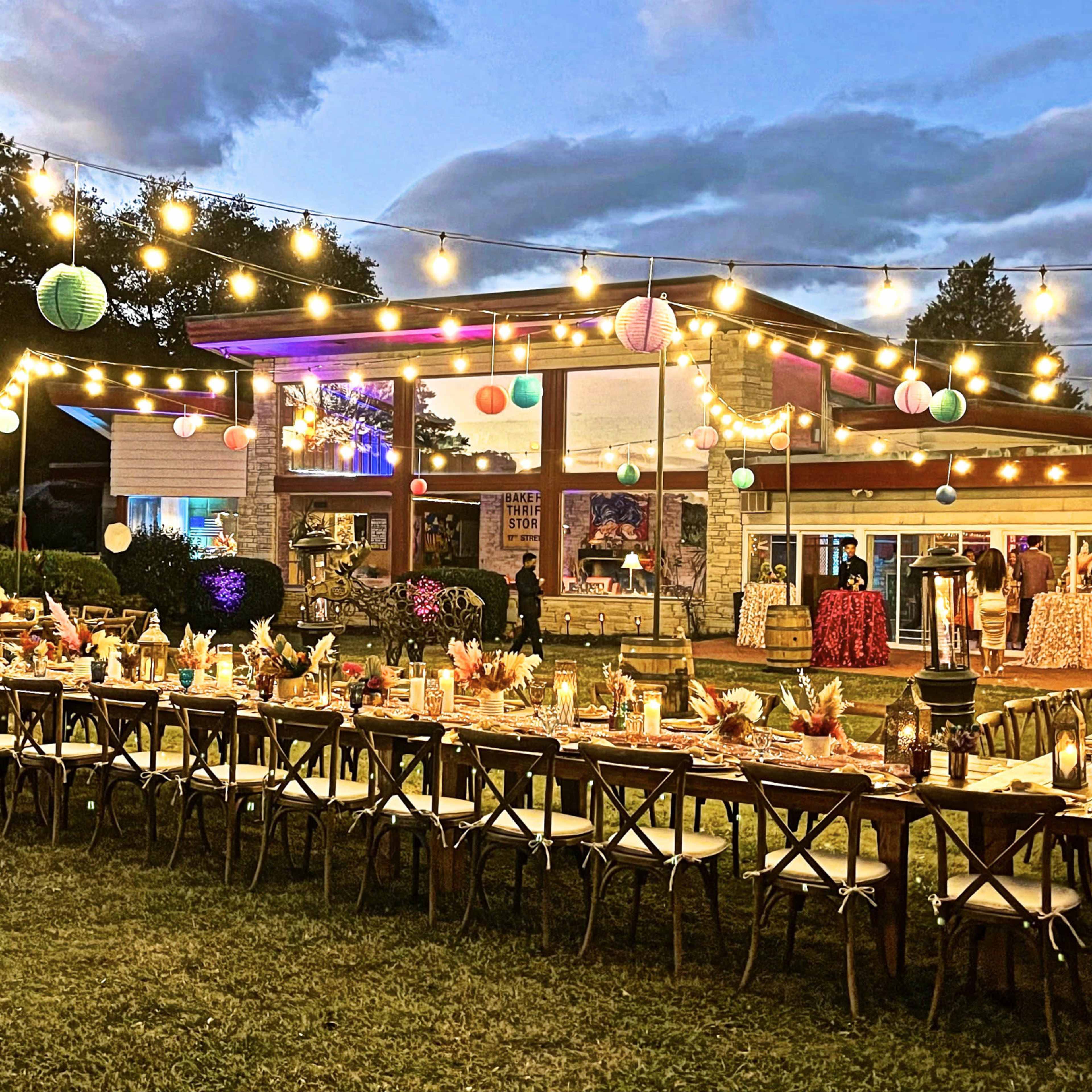 A large outdoor dining setup with a long table adorned with colorful decorations and lights, situated next to a modern building under a twilight sky.