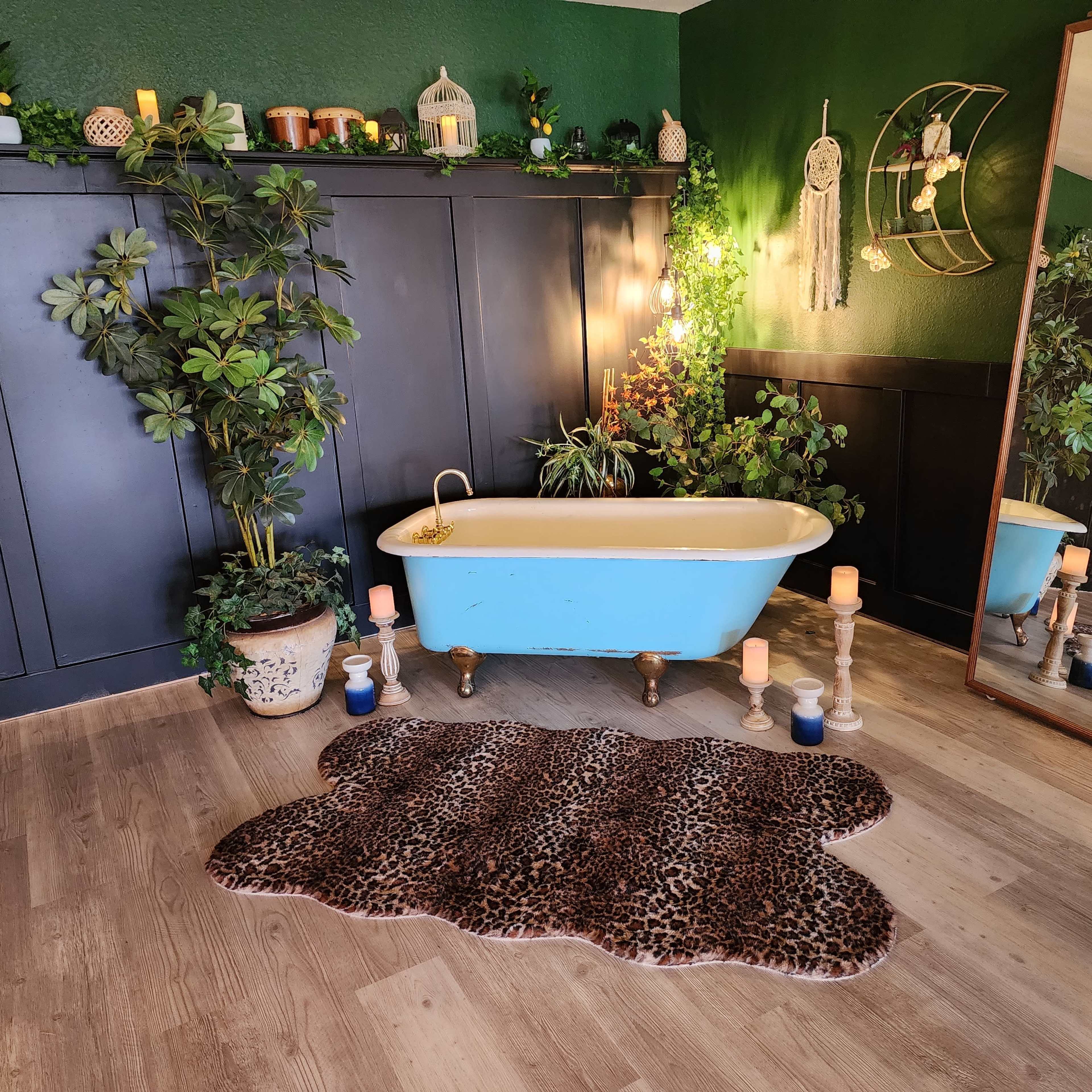 A vintage-style bathroom with a freestanding bathtub, surrounded by plants, candles, and a large mirror against a dark green wall.