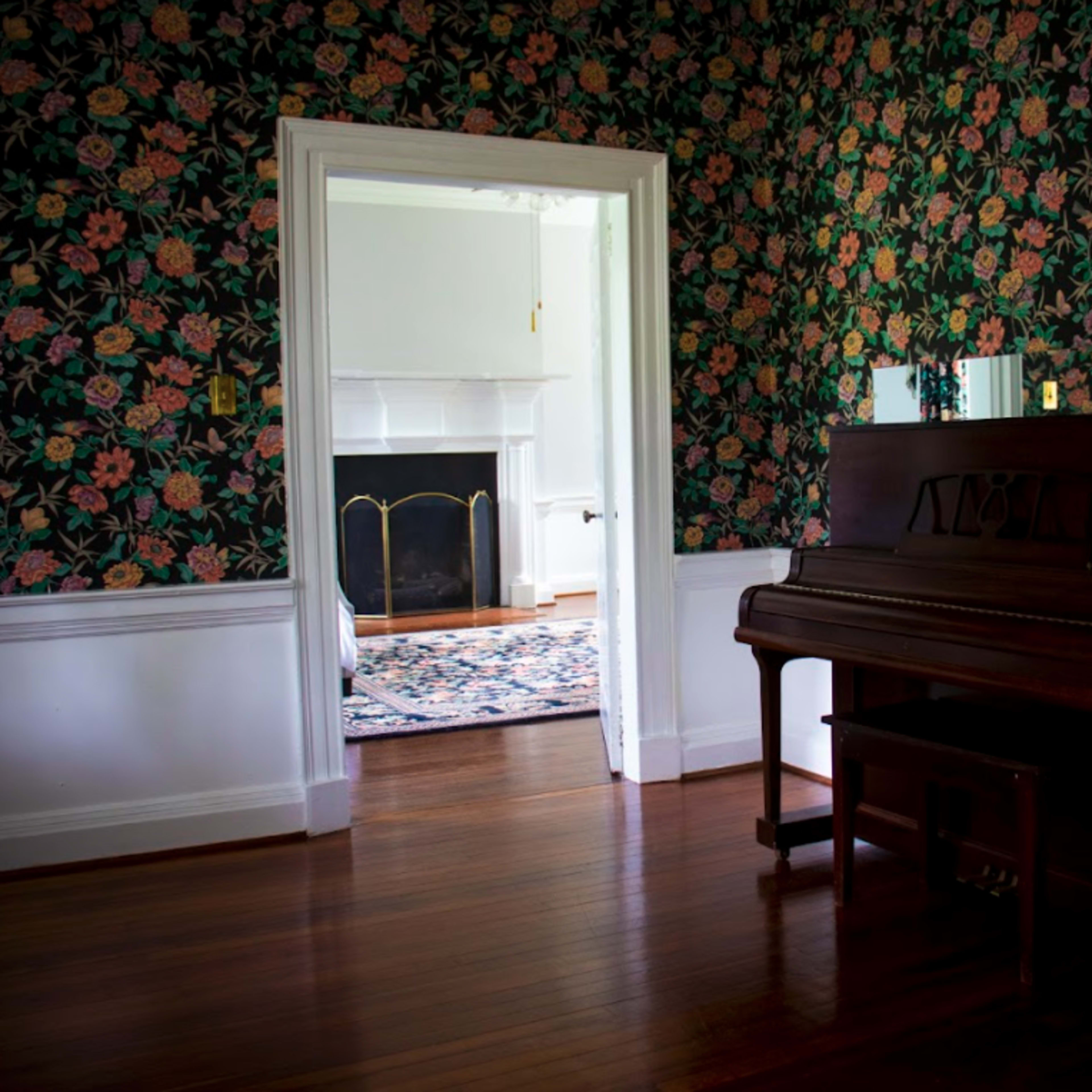 A wooden piano stands next to a floral-patterned wallpaper in a room that opens into a space with a fireplace and a visible rug.